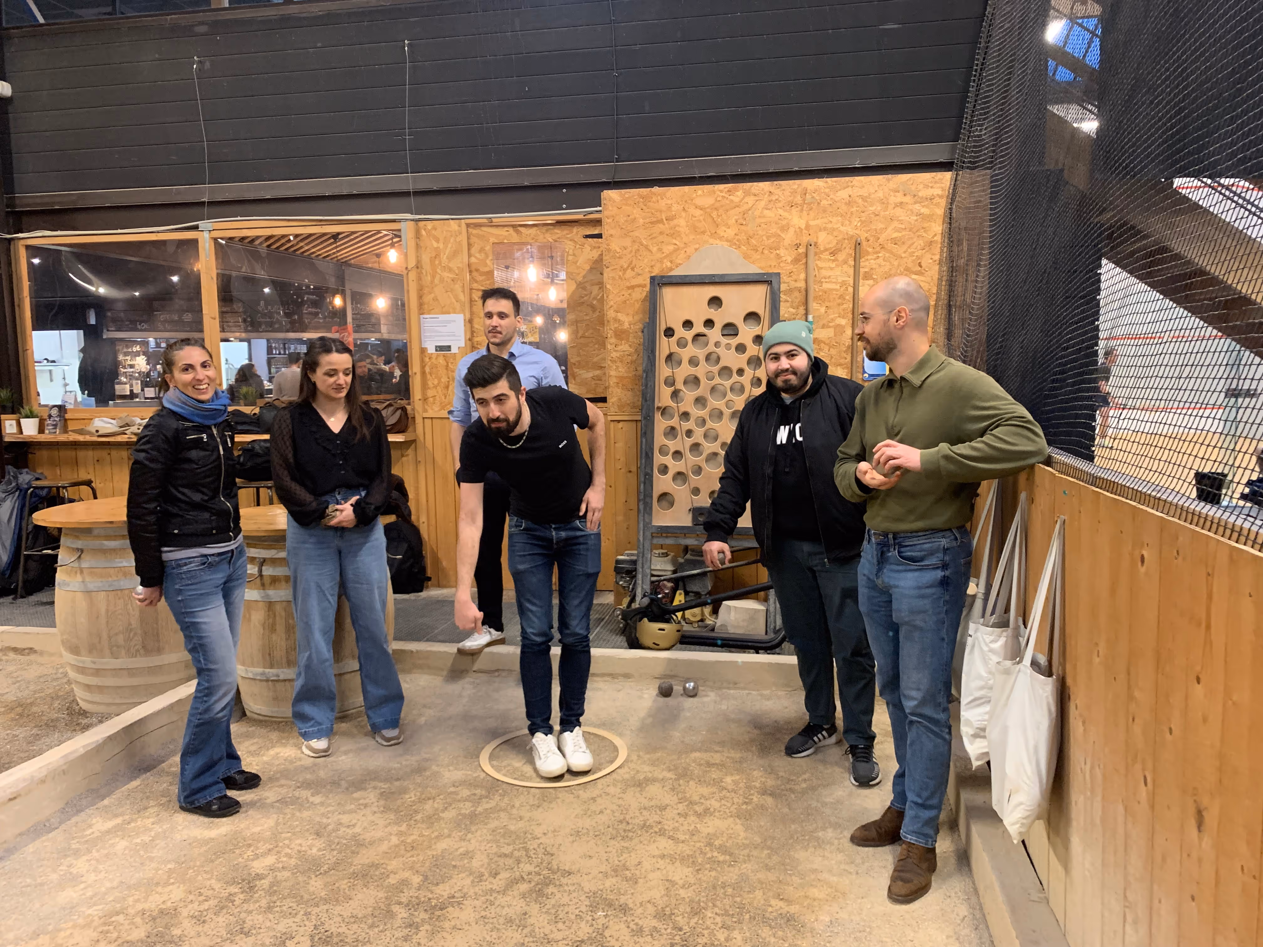 Groupe de six personnes jouant à la pétanque dans un espace intérieur avec un mur en bois et des tonneaux en arrière-plan.