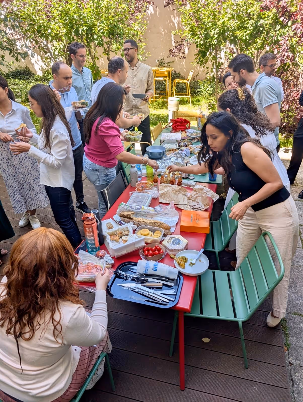 Un groupe de personnes profite d'un buffet en plein air avec diverses nourritures sur une table rouge dans un jardin.