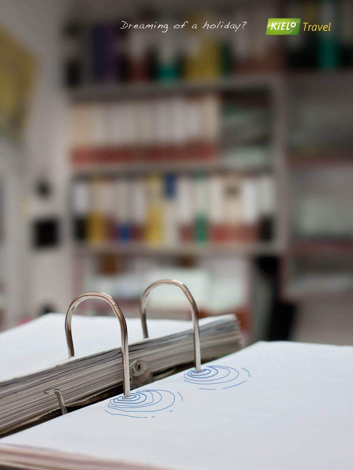 Kielo Travel print ad showing an office desk covered in paperwork and binders, with a turquoise holiday swimming pool doodled across one of the binders