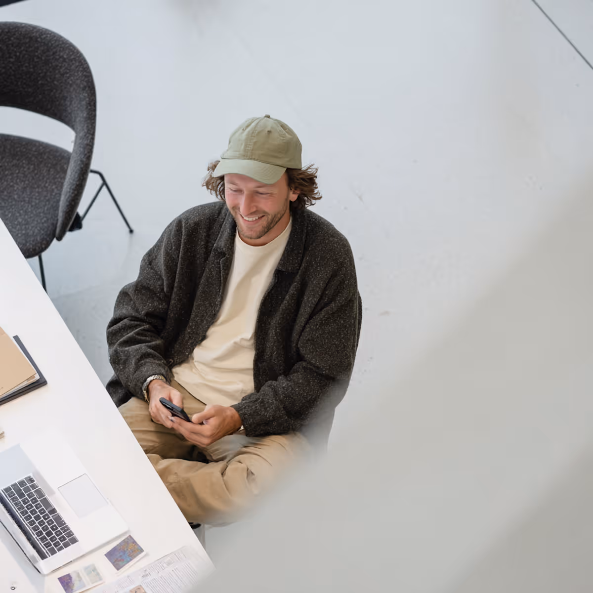 Man wearing cap and jacket sitting cross-legged at a desk smiling while holding a smartphone.
