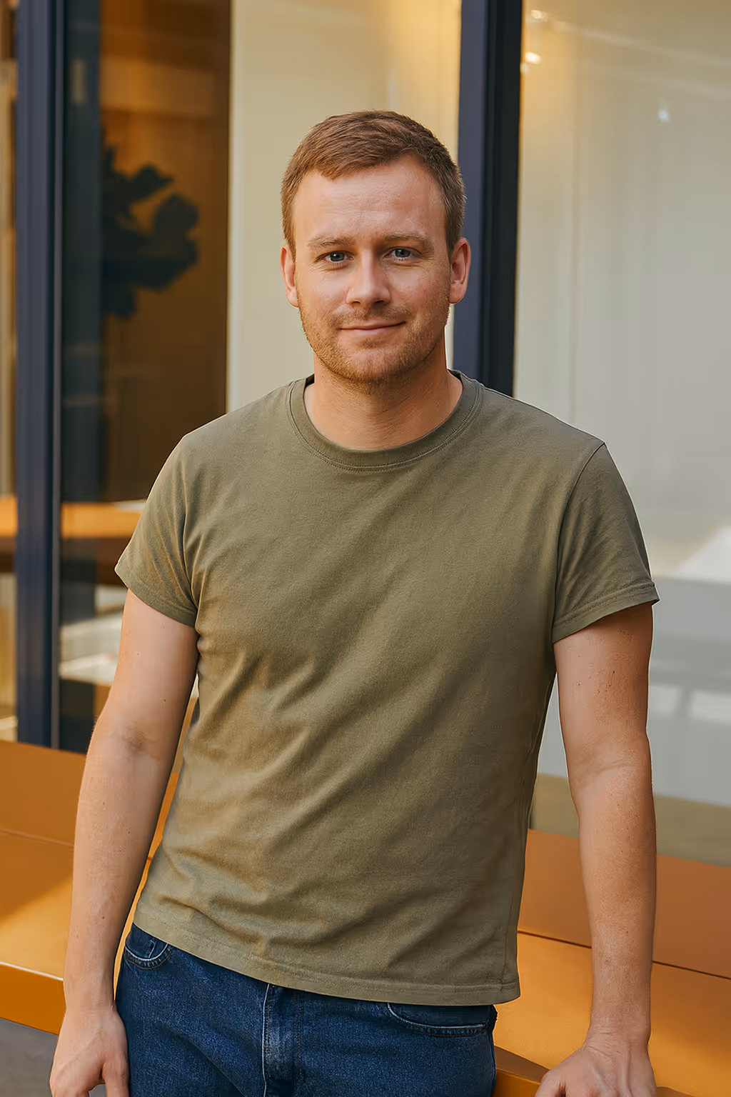Smiling man with short hair wearing an olive green t-shirt and blue jeans indoors.