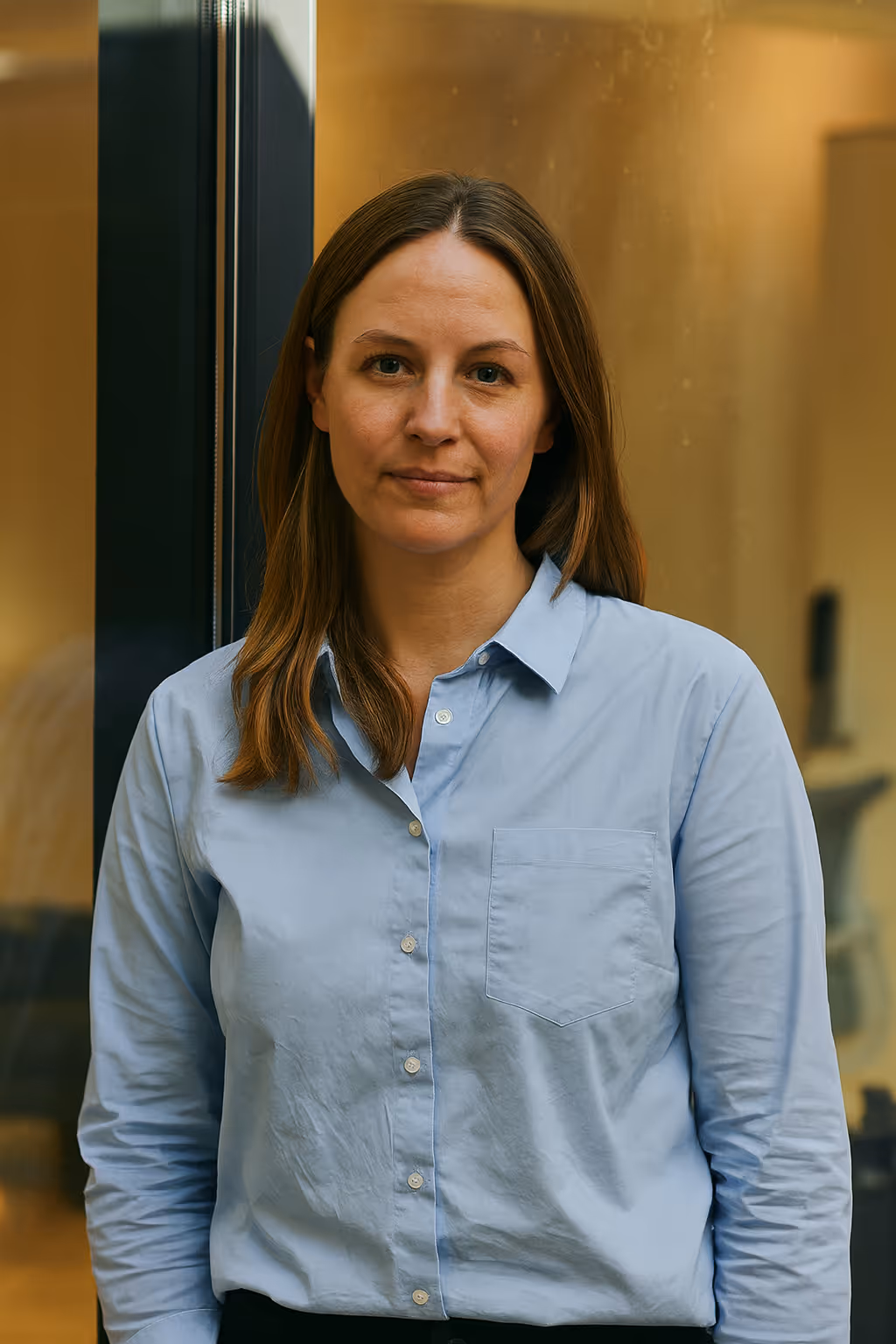 Woman with straight brown hair wearing a light blue button-up shirt standing indoors by a glass wall.