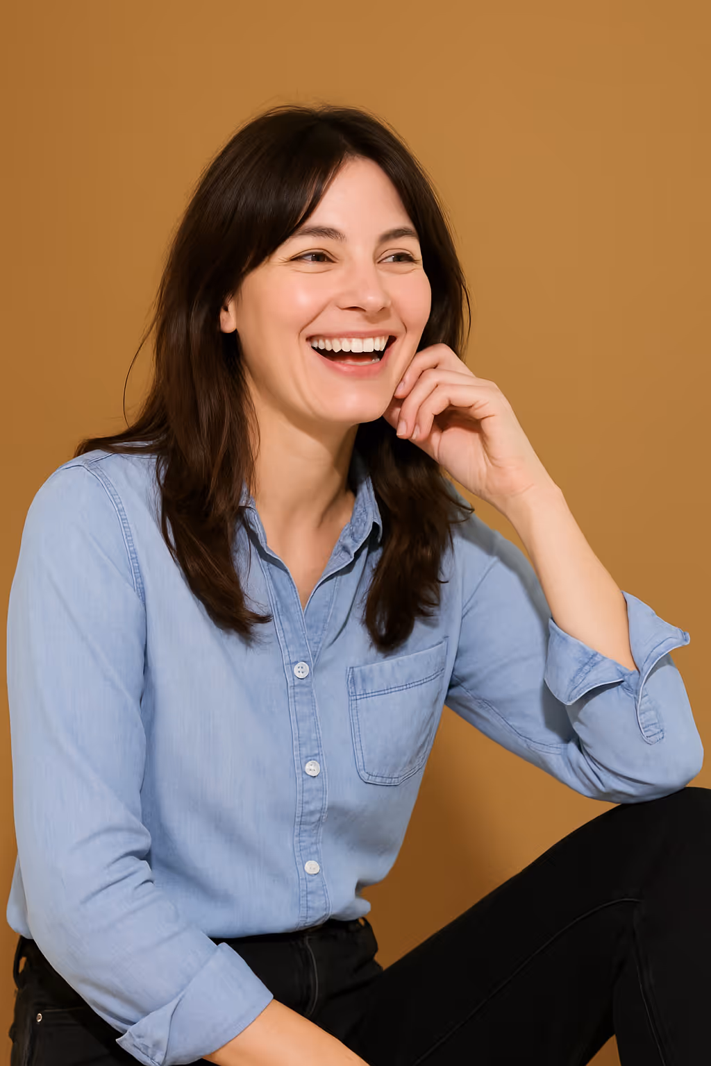 Smiling woman with dark hair in a light blue shirt sitting against a brown background.