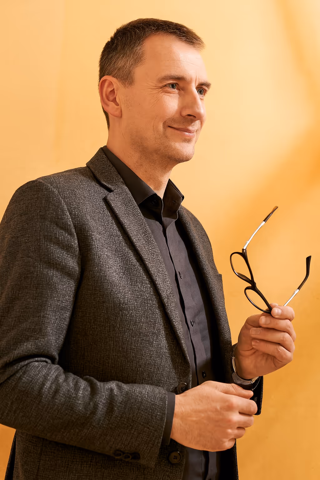 Smiling man in gray jacket holding black eyeglasses against a beige background.