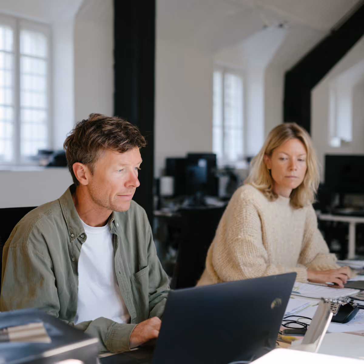 Man and woman working at a desk with laptops in a bright office with large windows.