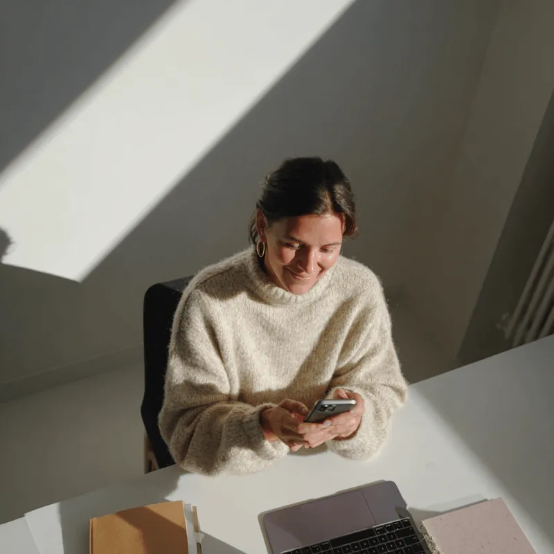 Woman in cream sweater smiling while using a smartphone at a desk with a laptop and notebooks.