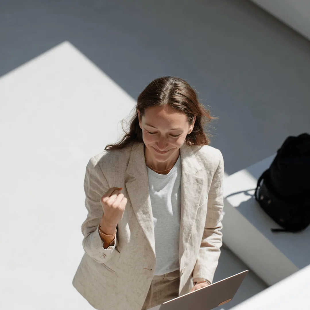 Woman in beige blazer celebrating with a fist pump while holding a laptop outdoors in sunlight.