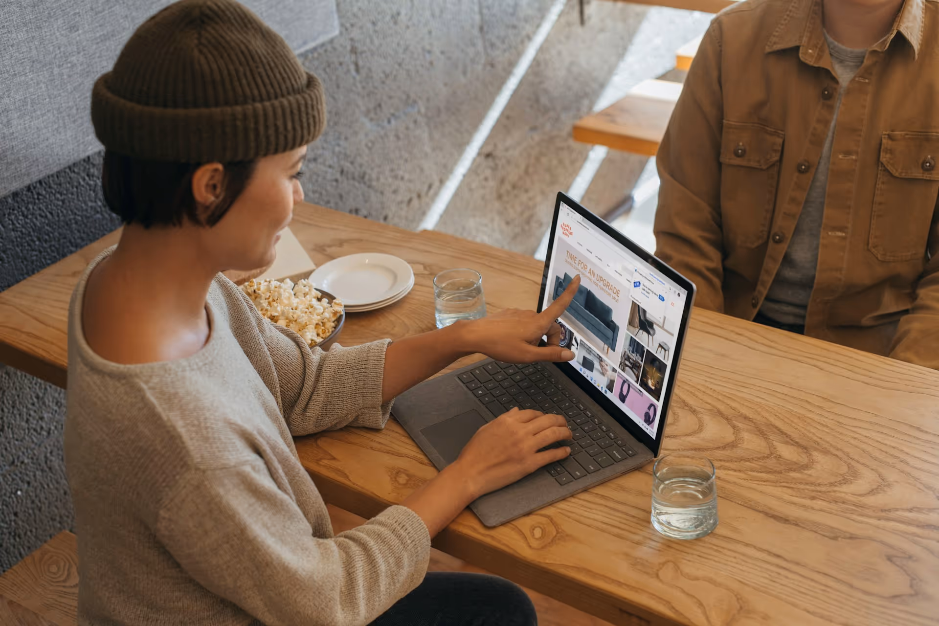 Person pointing at a laptop screen showing furniture and home decor while sitting at a wooden table.