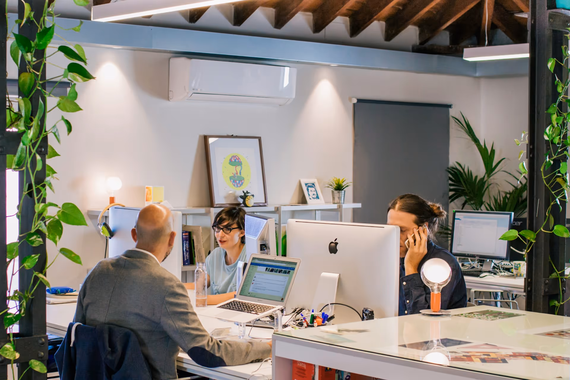 Three people working at desks with computers in a modern office with plants and artwork.