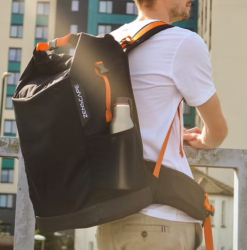 Person wearing a large black Zenscape backpack with orange straps, standing by a railing outdoors.