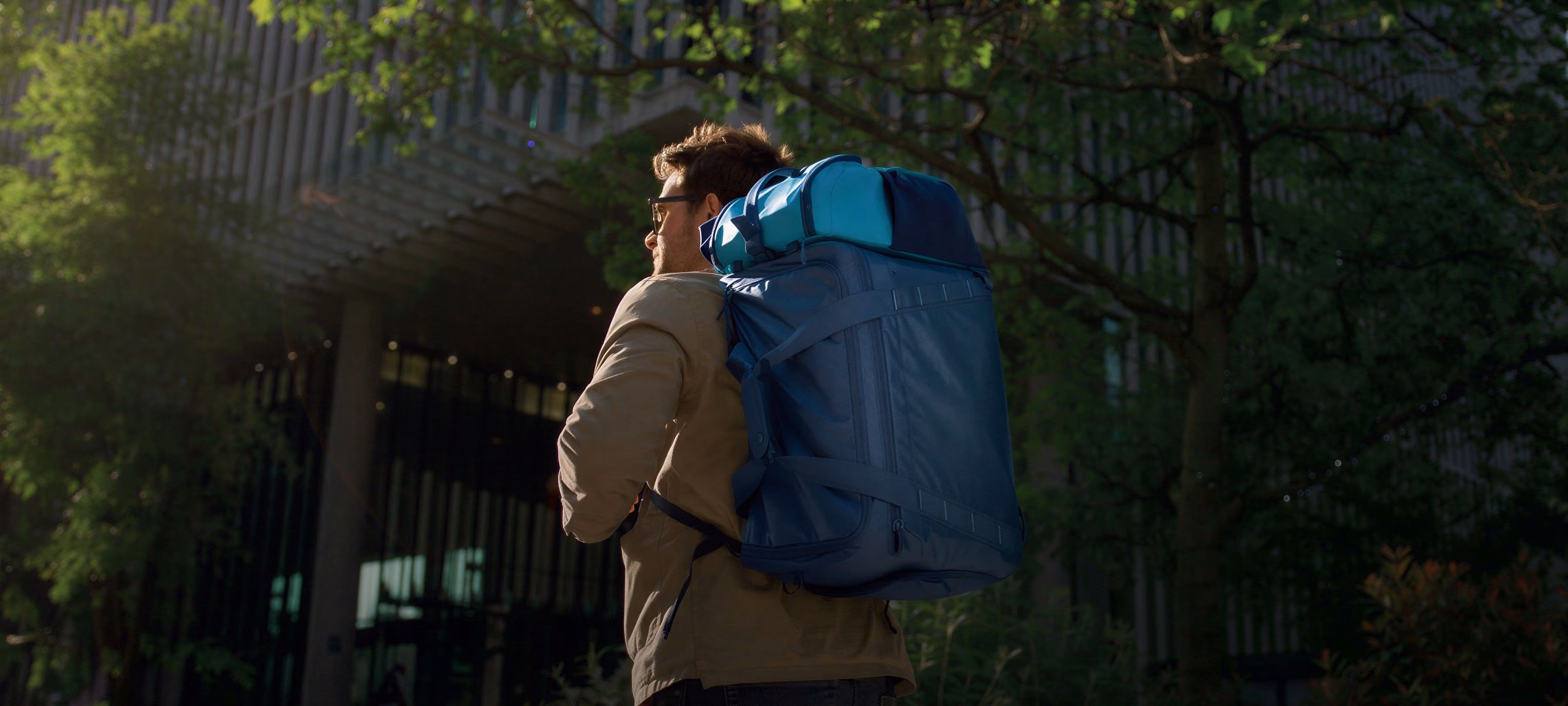 Man wearing sunglasses and carrying a large blue backpack walking outdoors near trees and a building.
