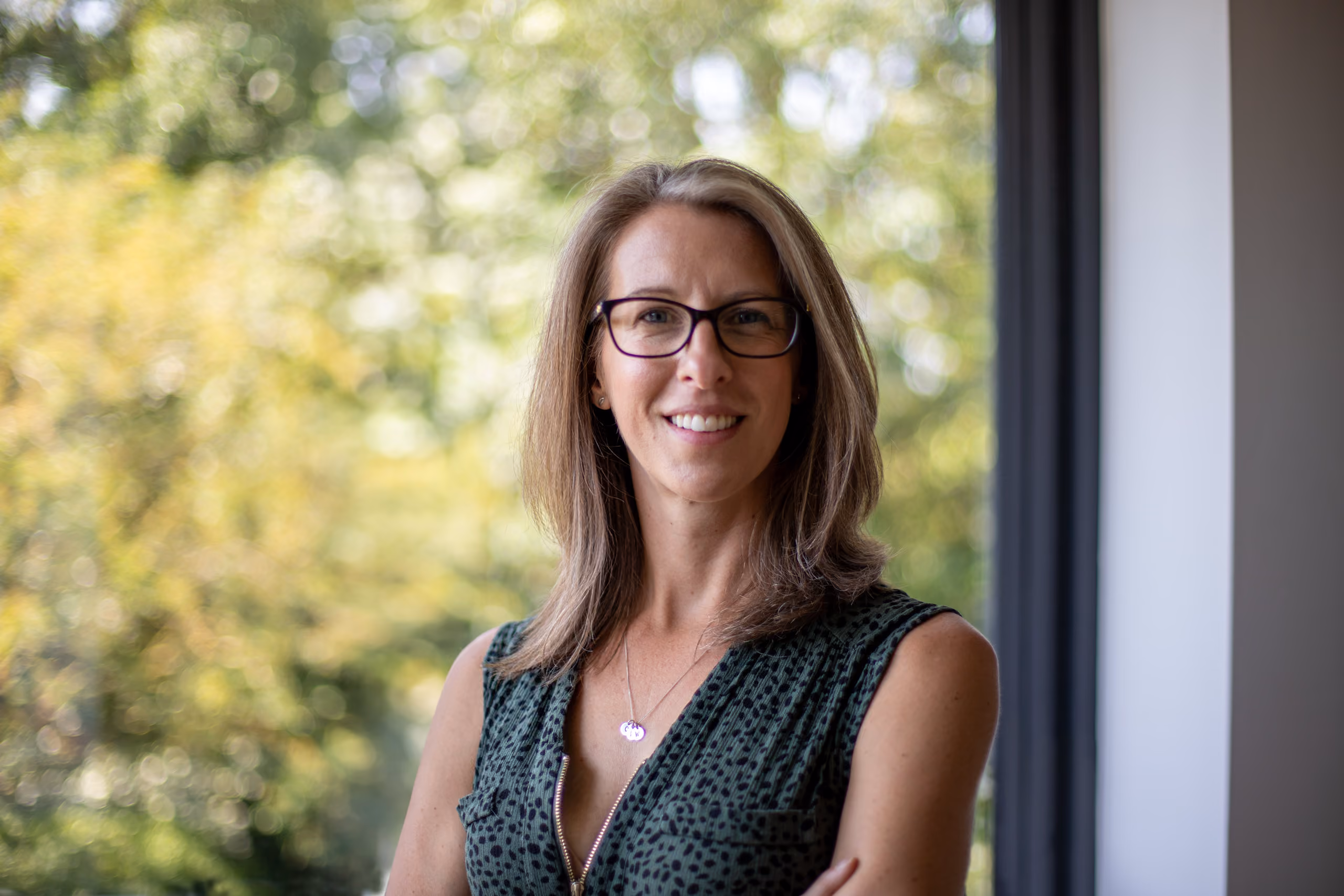 Smiling woman with glasses and shoulder-length hair standing by a window with greenery outside.