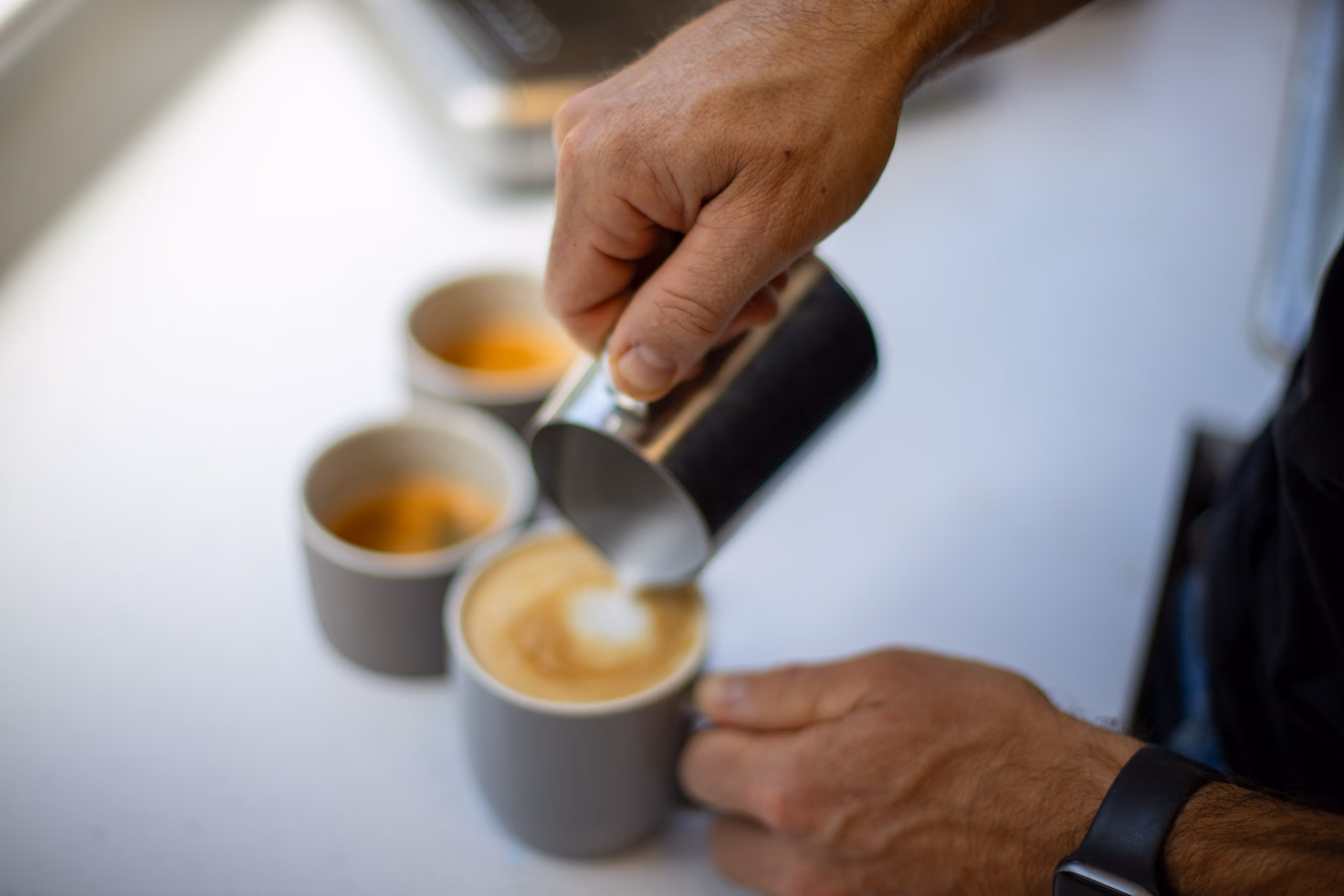 Hands pouring steamed milk from metal pitcher into a coffee cup to make latte art.