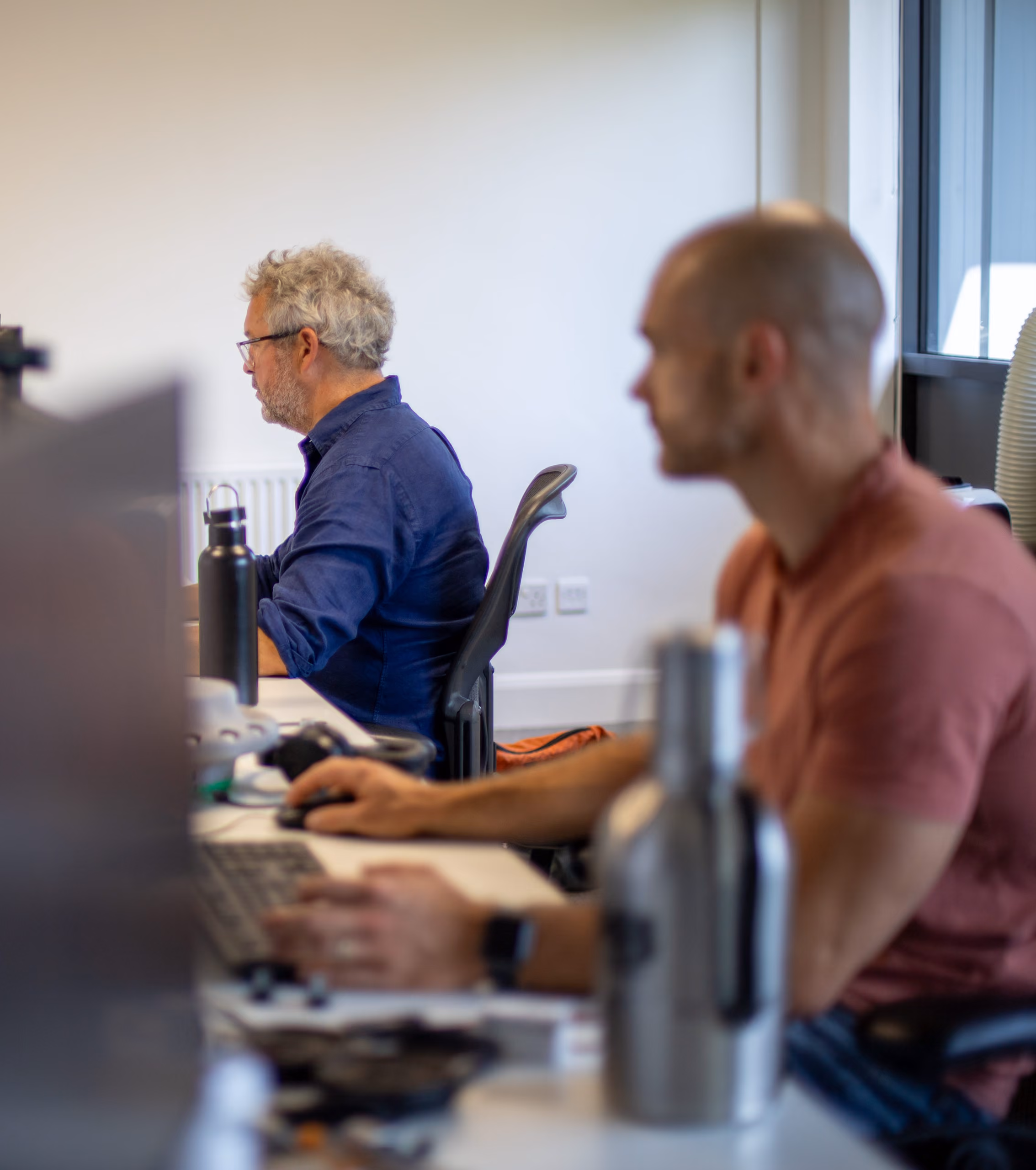 Two men working at computers in an office with water bottles on their desks.