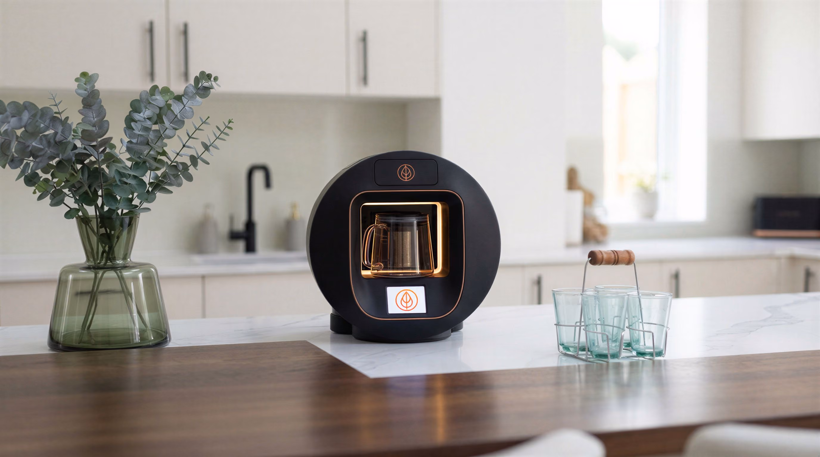 Black circular tea maker with a glass pot on a kitchen island next to a green vase and glass cup holder.