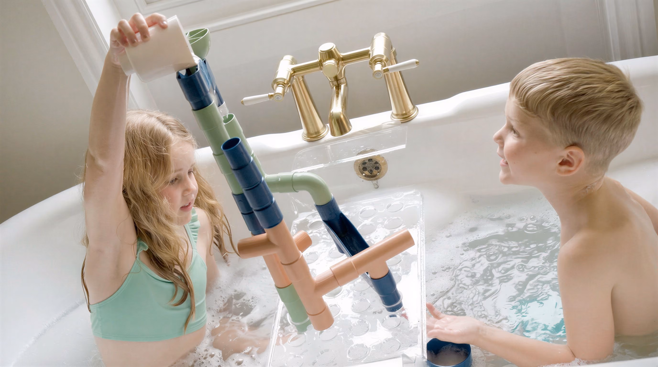 Two children in a bathtub playing with a colorful pipe water toy, girl pouring water from a cup.