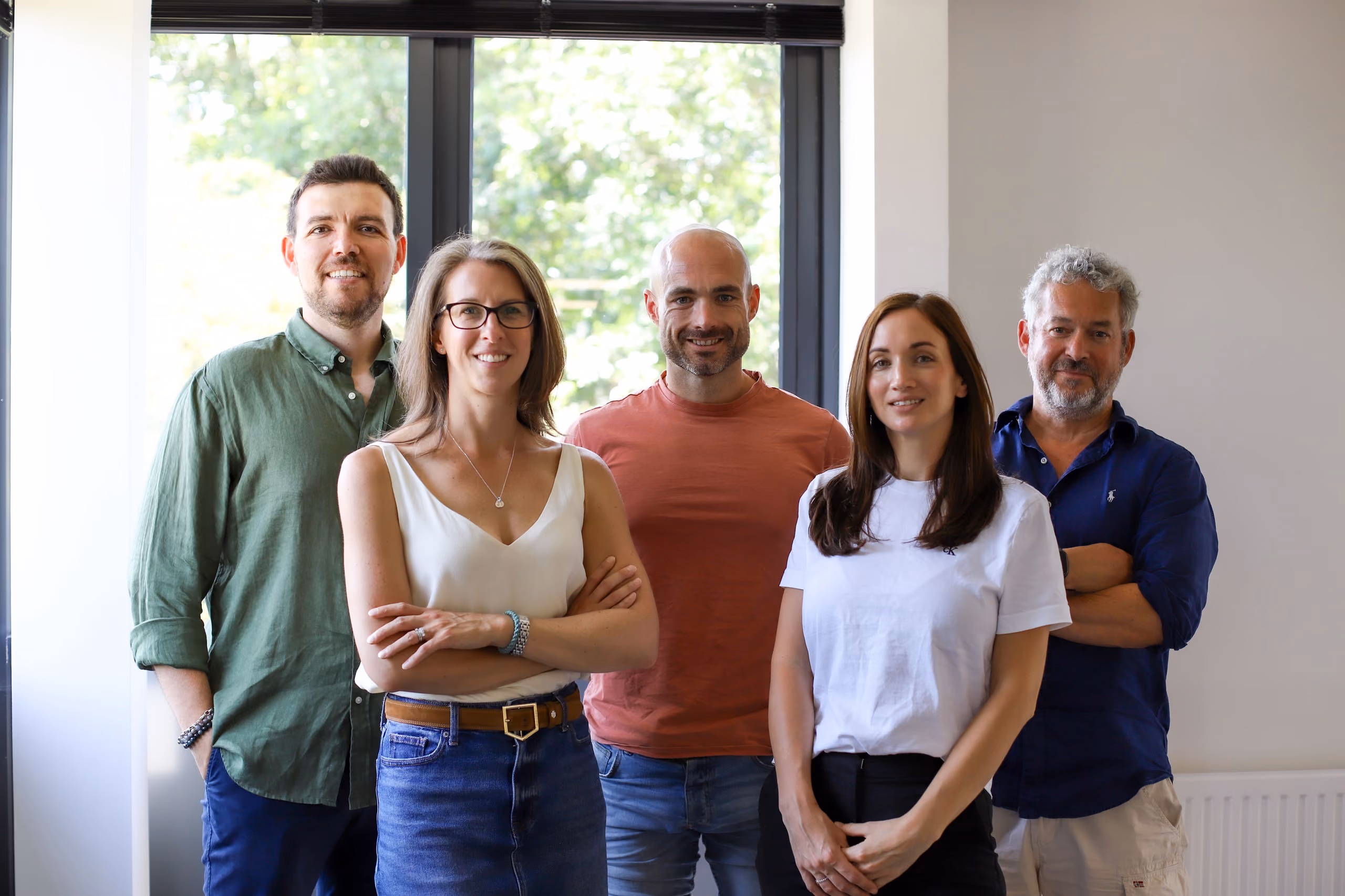 Group of five adults standing indoors near a window, smiling and facing the camera.