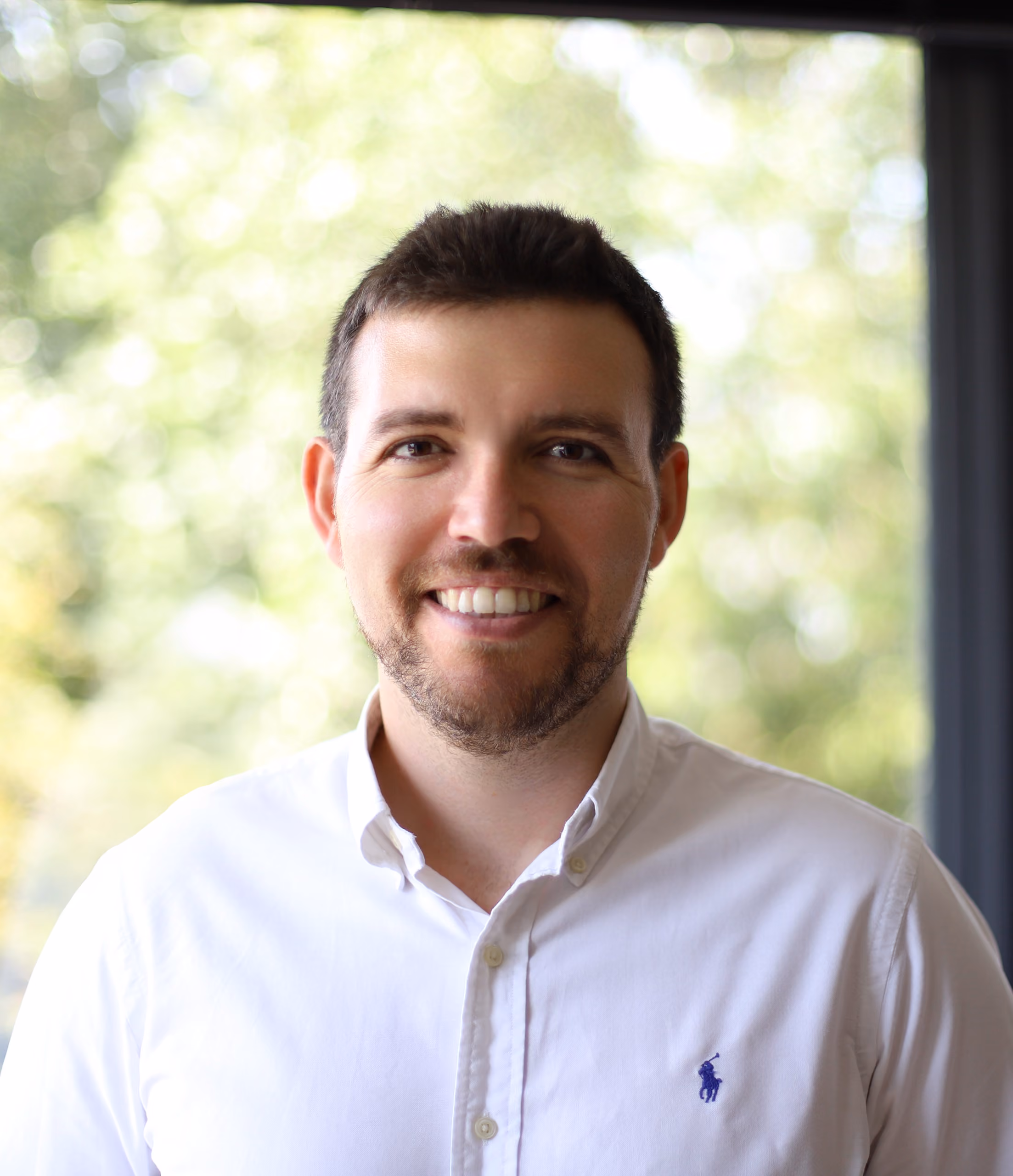 Smiling man with short dark hair and beard wearing a white button-up shirt by a bright window.