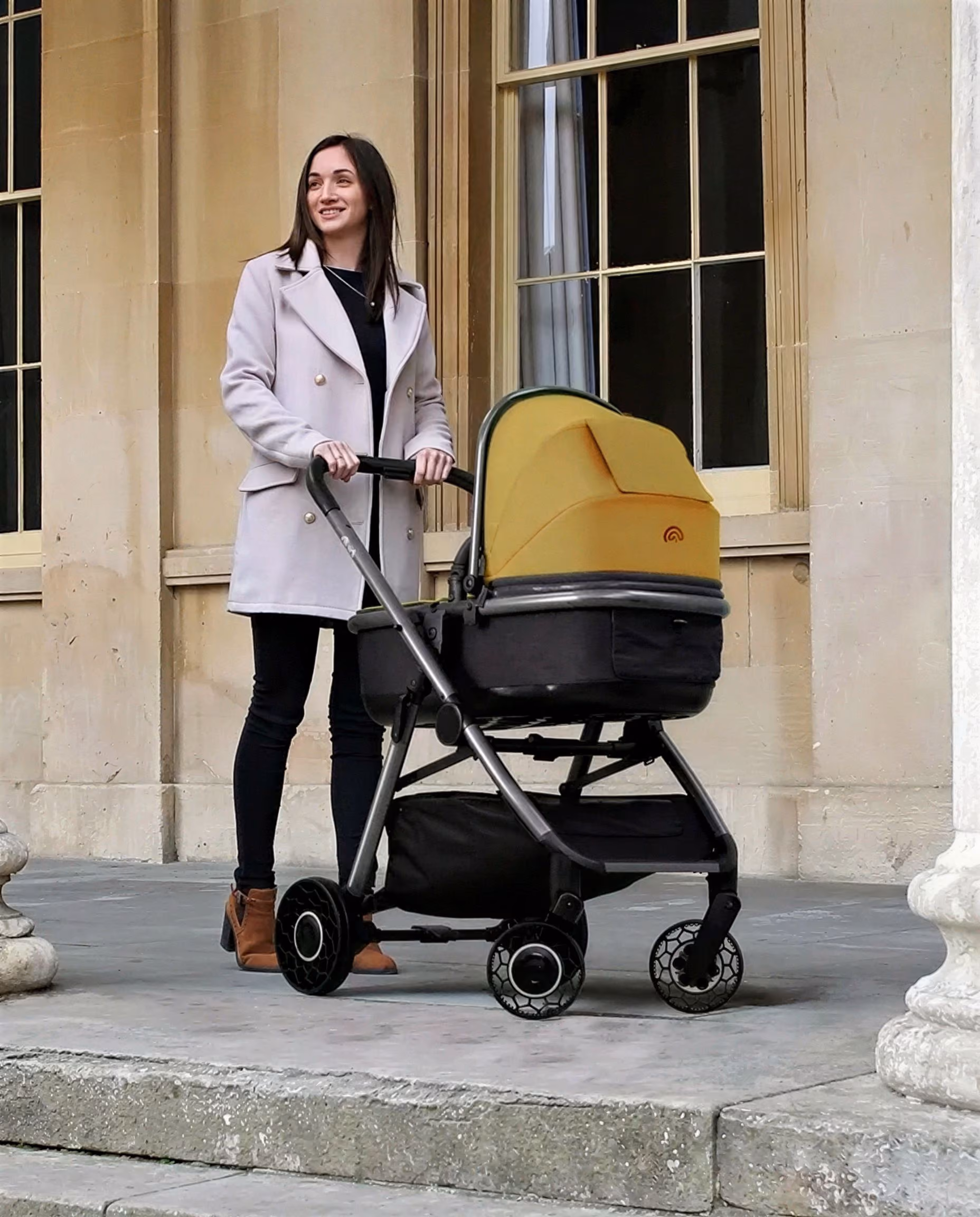 Woman in light coat pushing a black and yellow baby stroller on stone porch in front of windows.