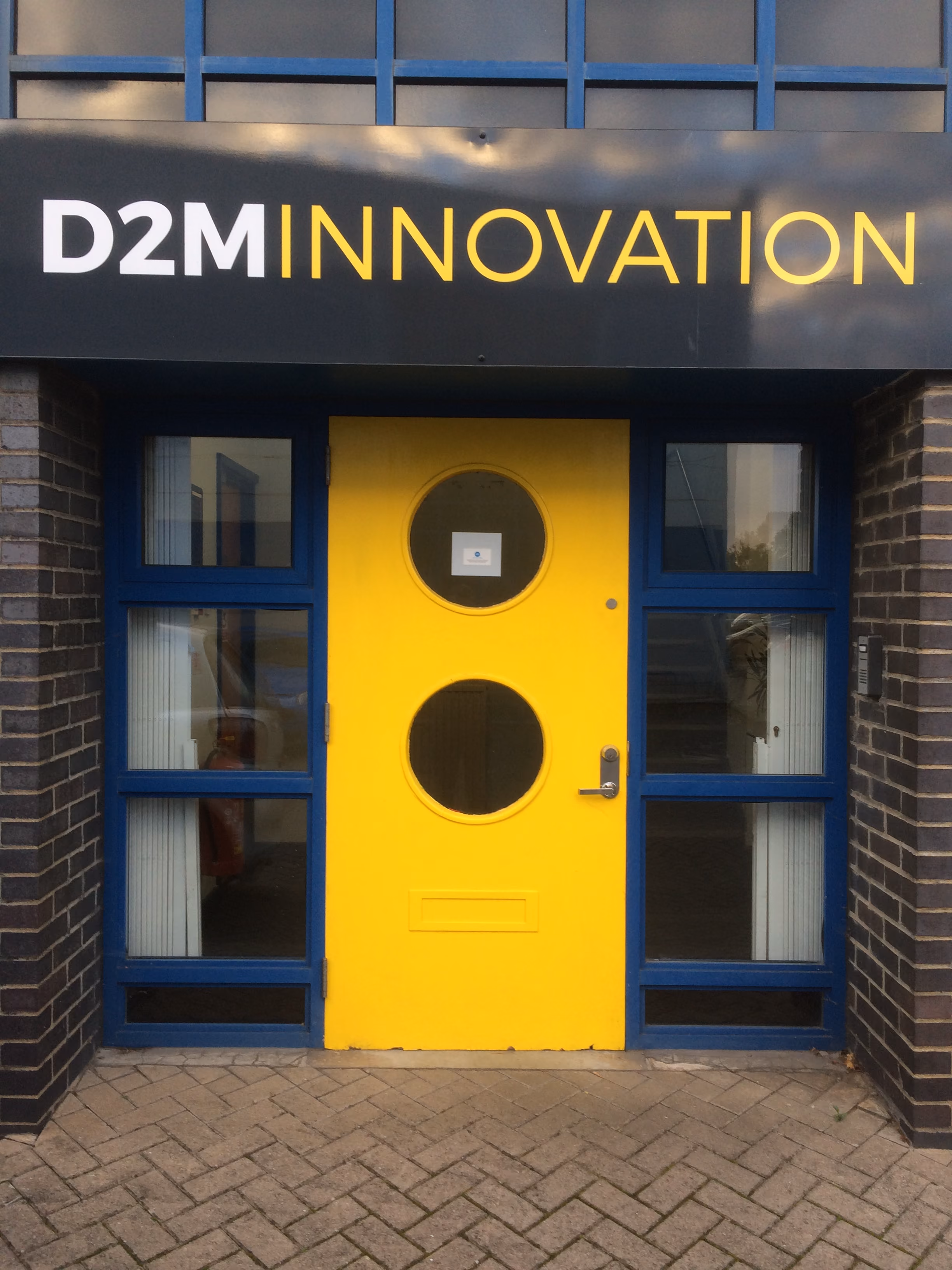 Bright yellow door with two round windows set in a blue-framed entrance under a sign reading D2M Innovation.