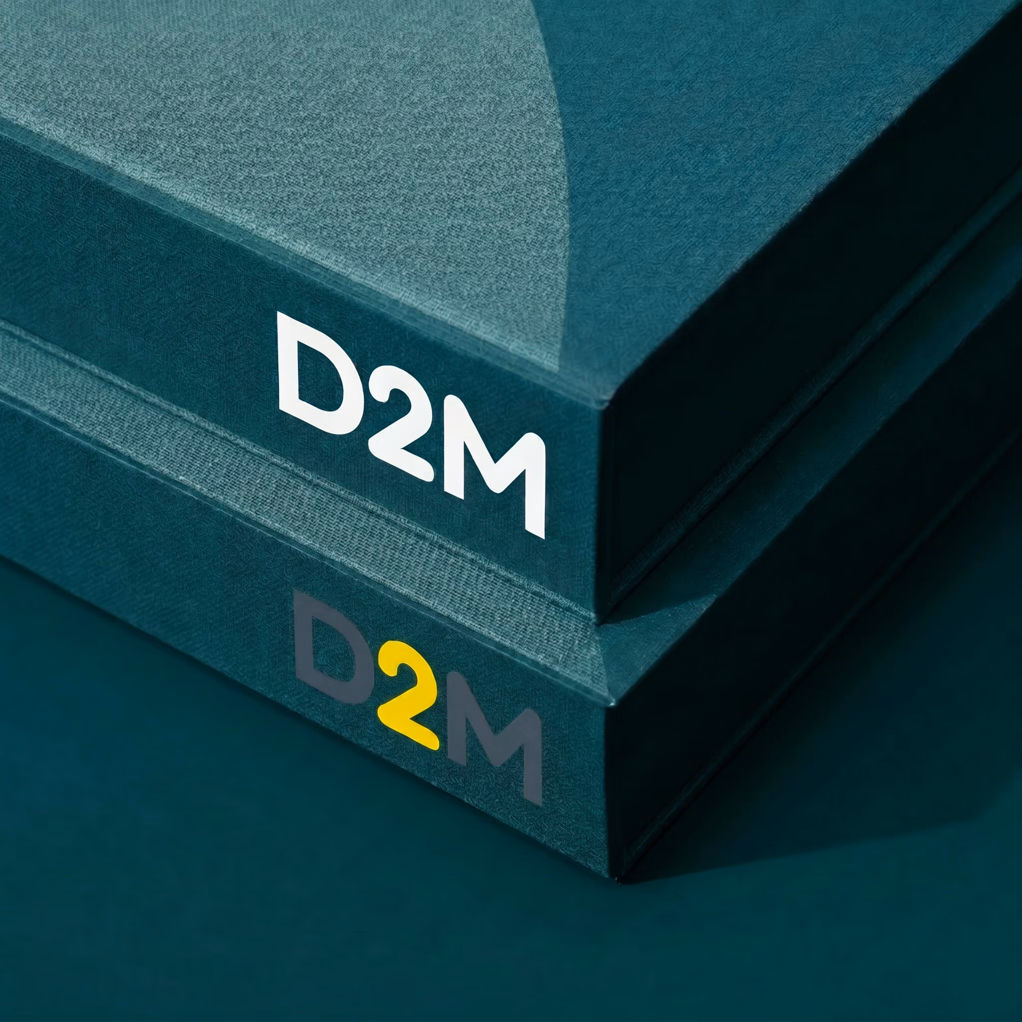 Two stacked dark teal books with the text D2M on their spines, one in white and one in gray and yellow.