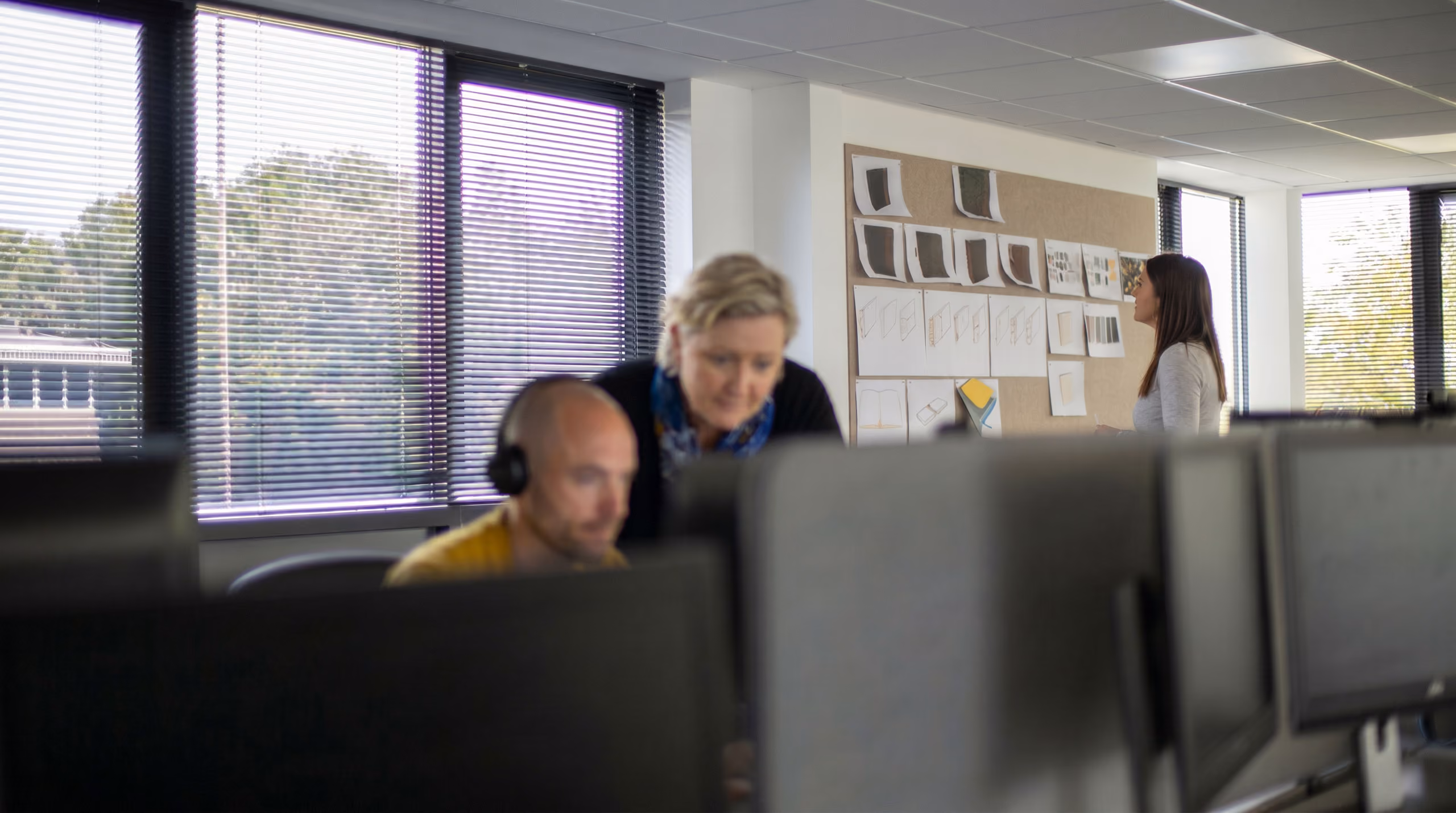 Office scene with a man wearing headset at a desk, woman leaning over him and another woman looking at pinned papers.