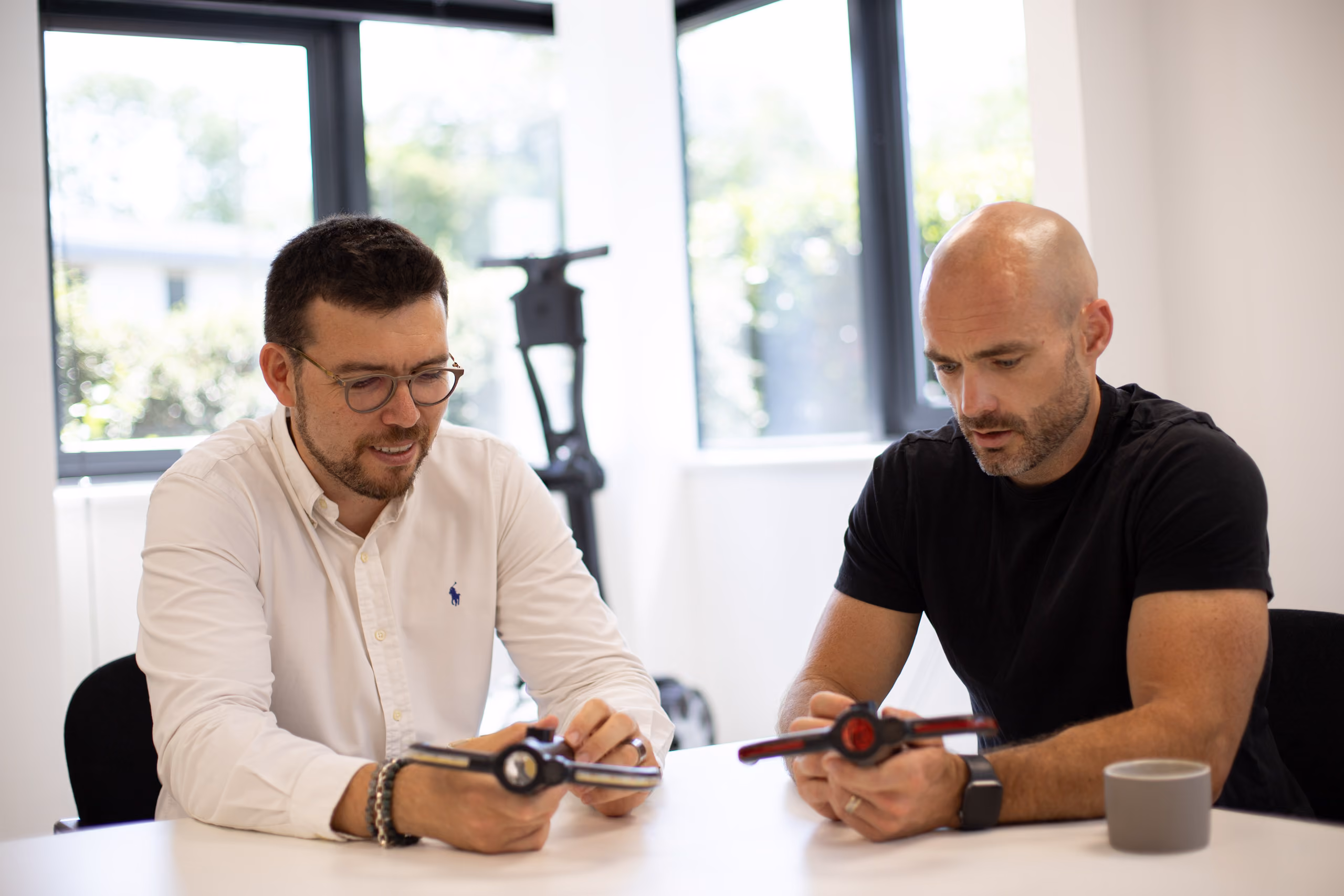 Two men sitting at a table examining black and red mechanical parts in a bright room.