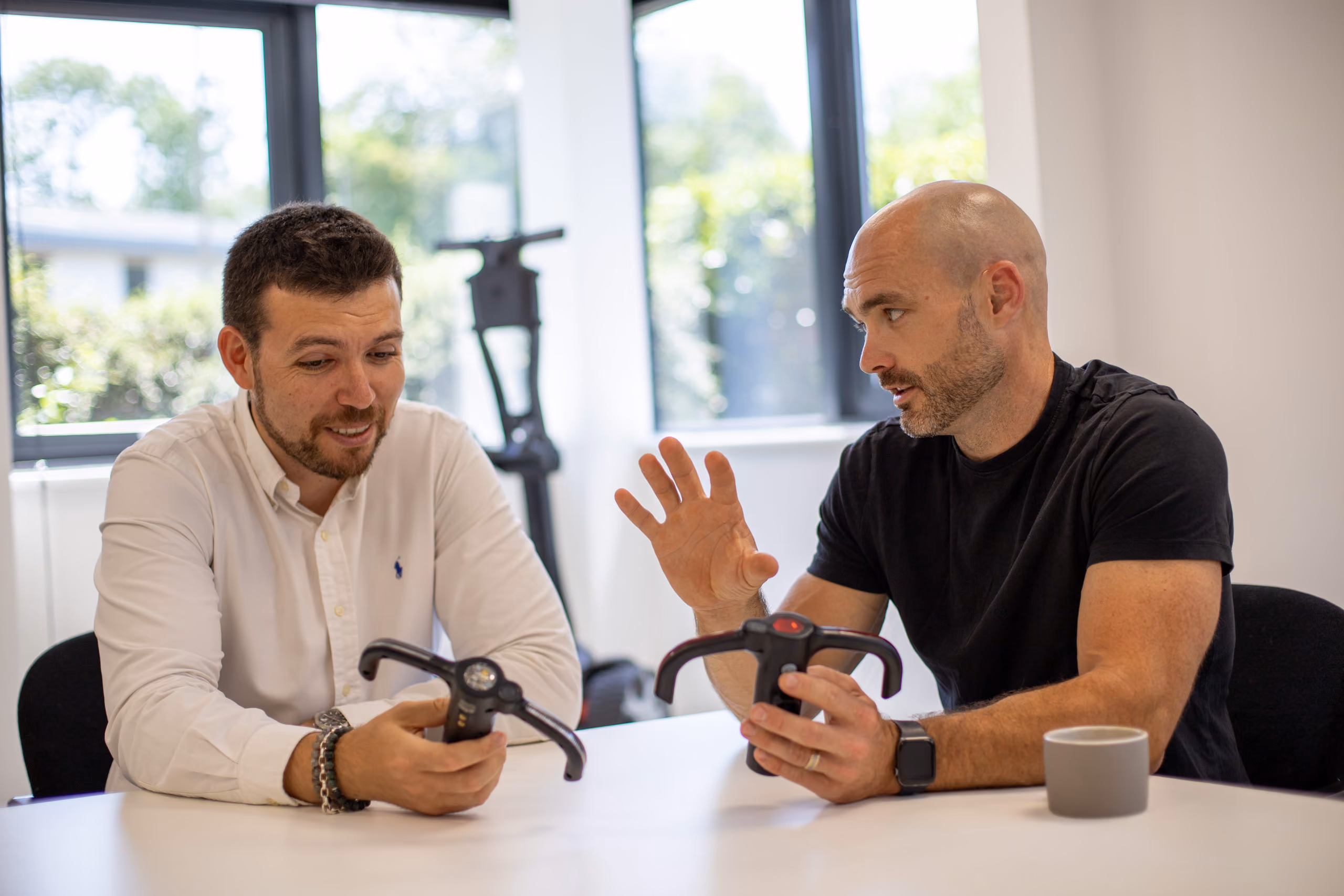 Two men sit at a table holding and discussing electric scooter handlebars in a bright room.