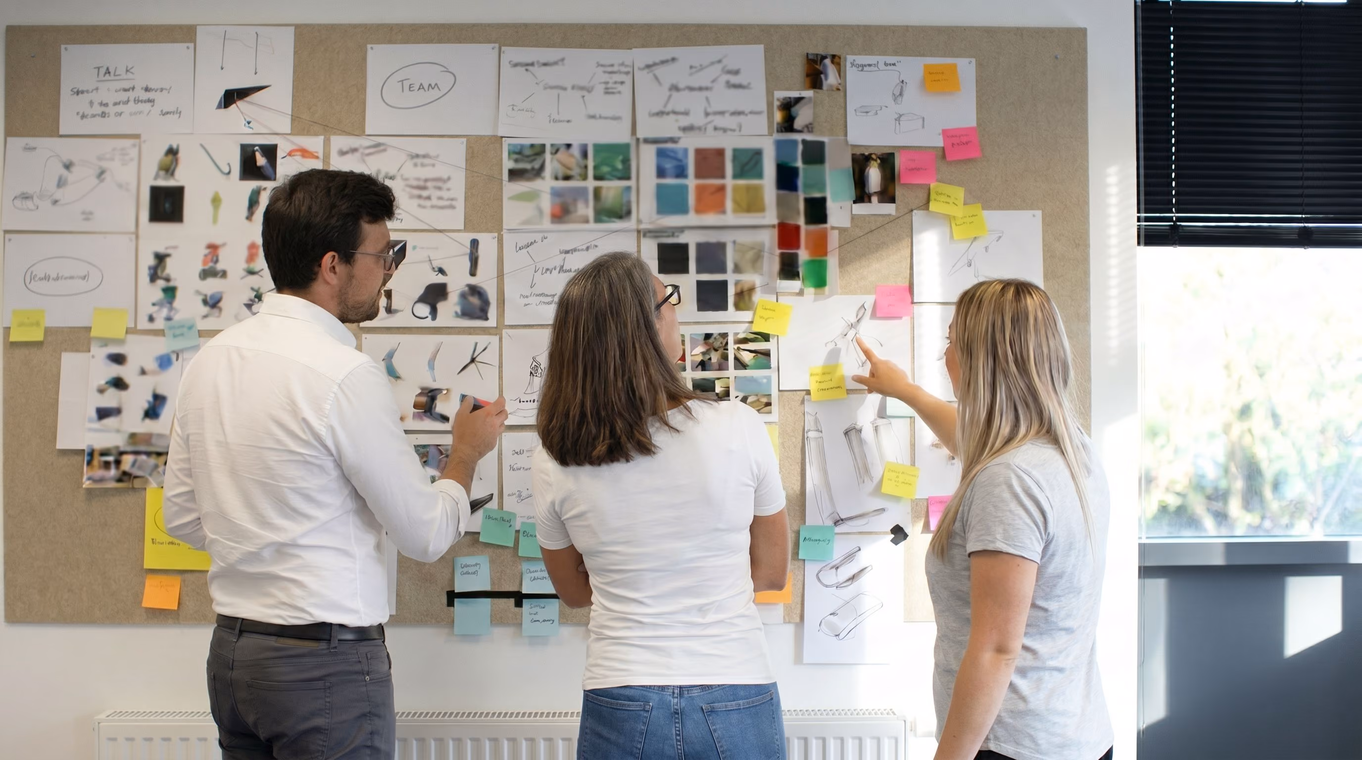 Three people reviewing design sketches and notes pinned on a board in a bright room.