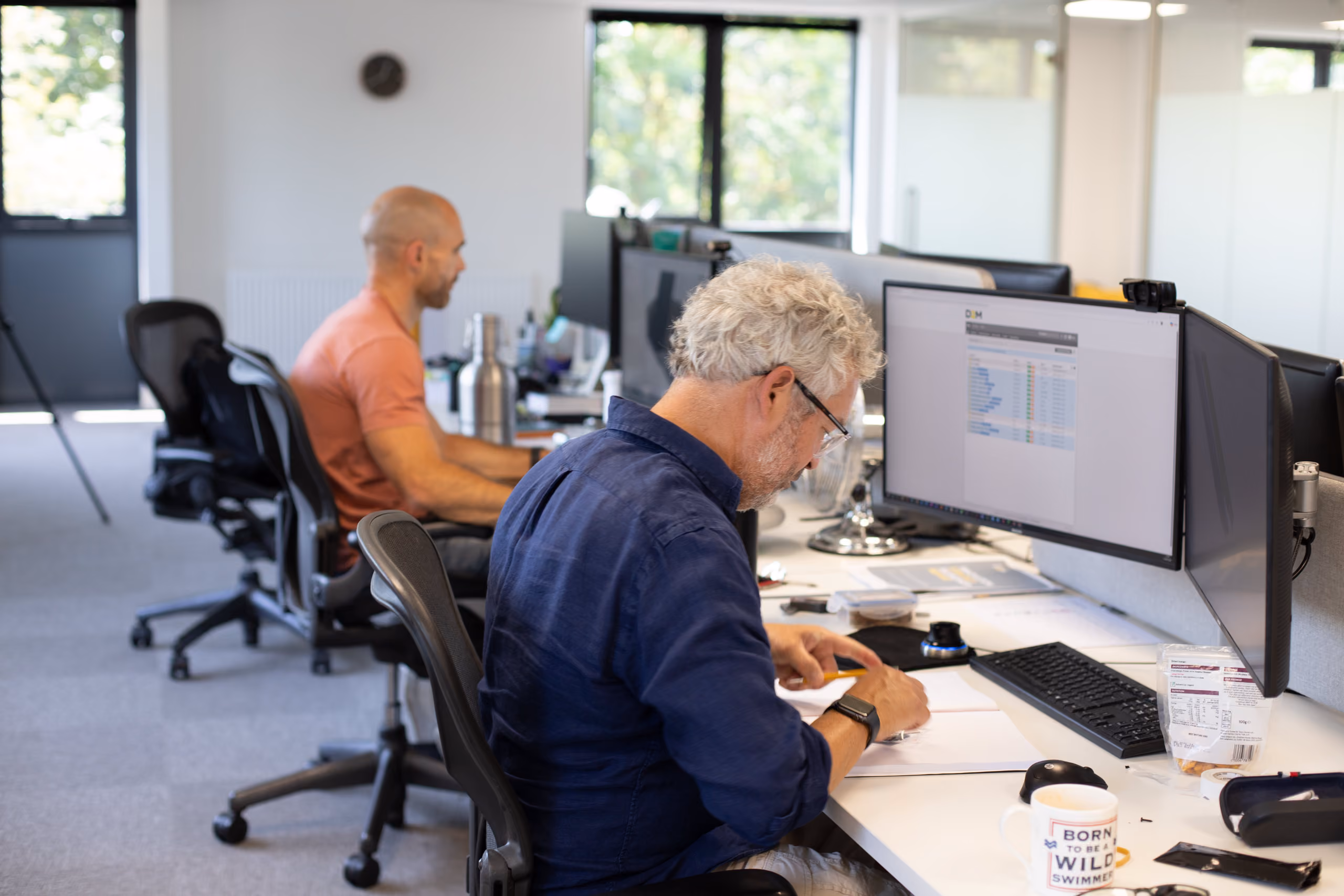 Two men working at desks in an office, one using a computer, the other writing on paper.
