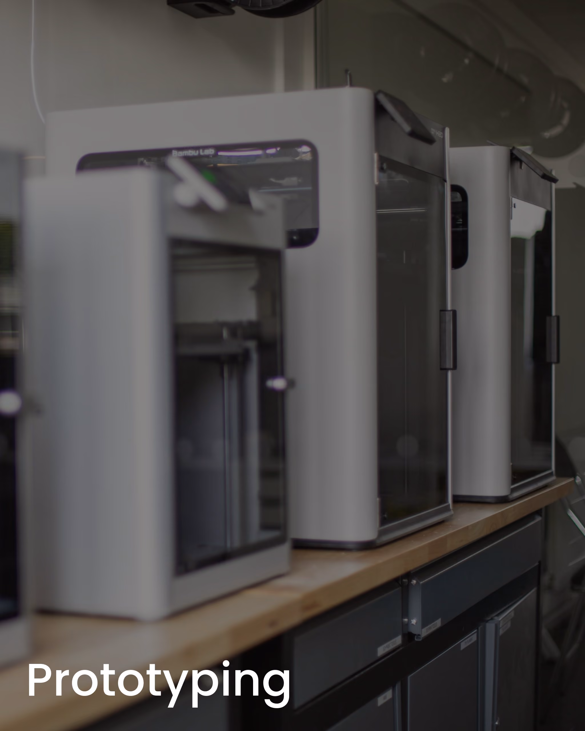 Row of white 3D printers on a wooden table in a prototyping lab.