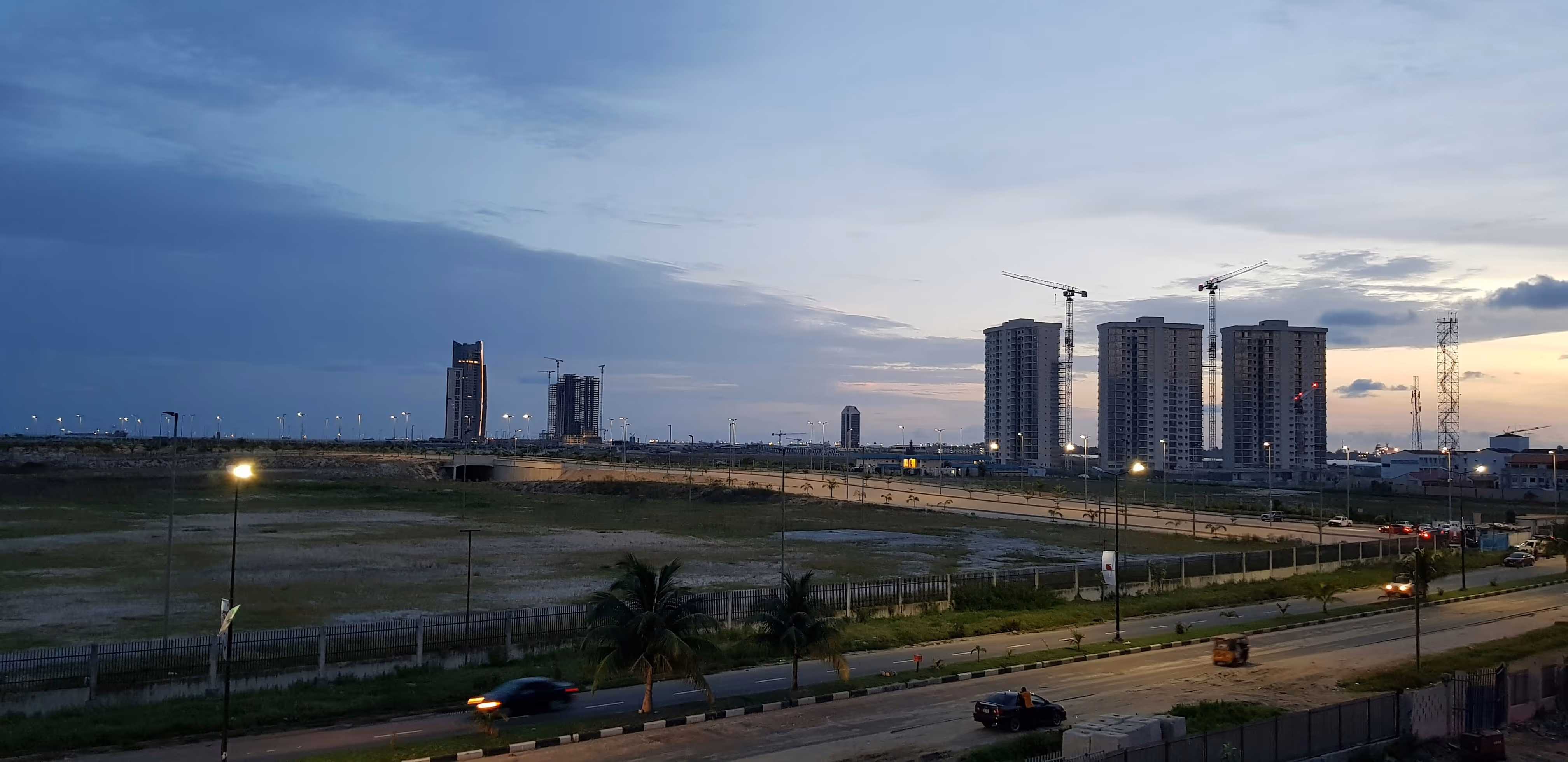 Dusk cityscape showing tall buildings under construction with cranes, streetlights on, vehicles on roads, and a partly cloudy sky.