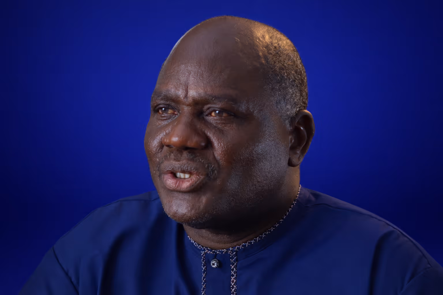 Portrait of an older man with dark skin wearing a blue shirt, set against a blue background.