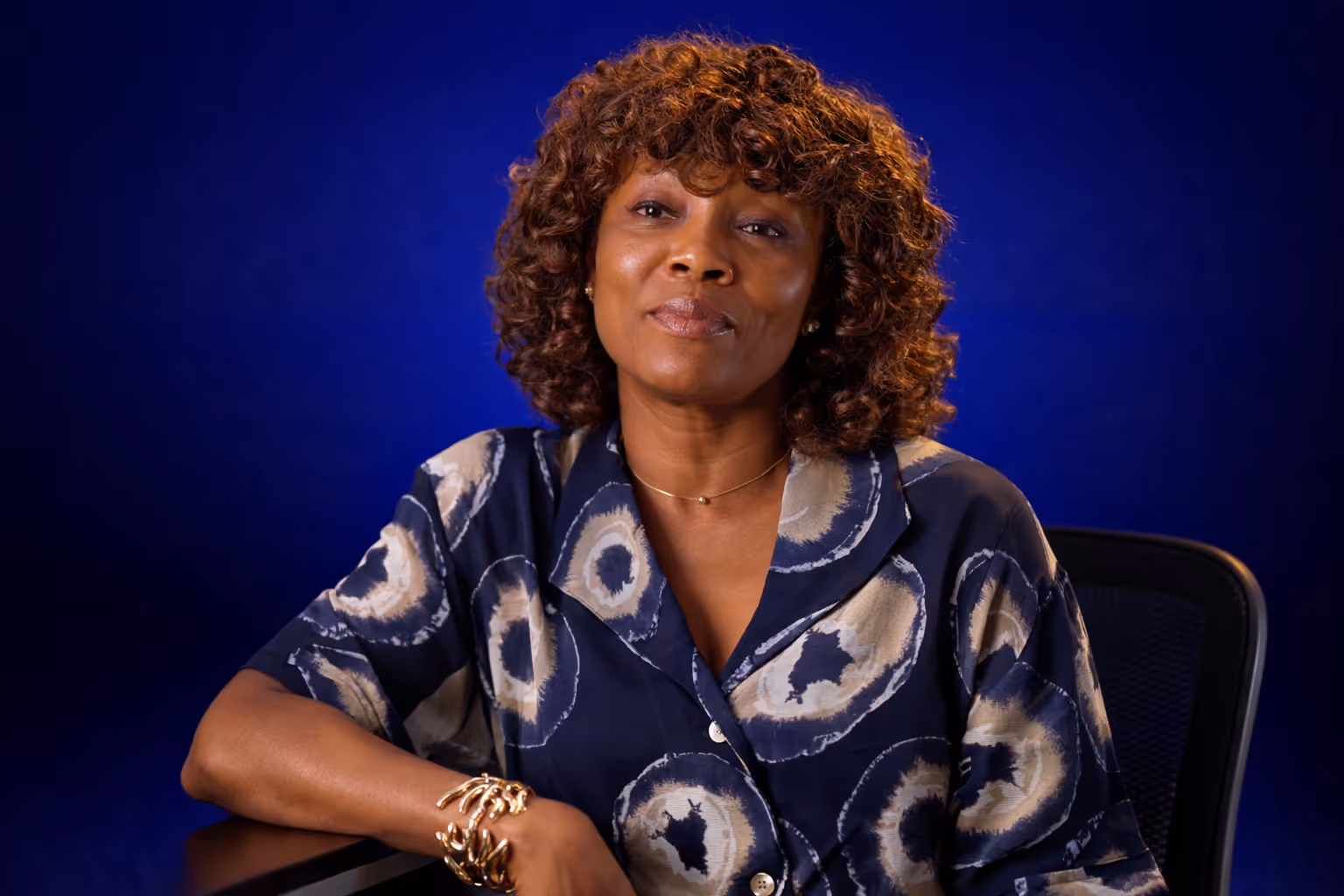 Woman with curly hair wearing a patterned navy blue blouse and gold bracelet sitting with arm resting on a table against a blue background.
