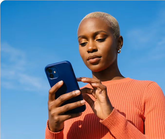 Woman with short blonde hair wearing an orange sweater looking at a smartphone against a clear blue sky.