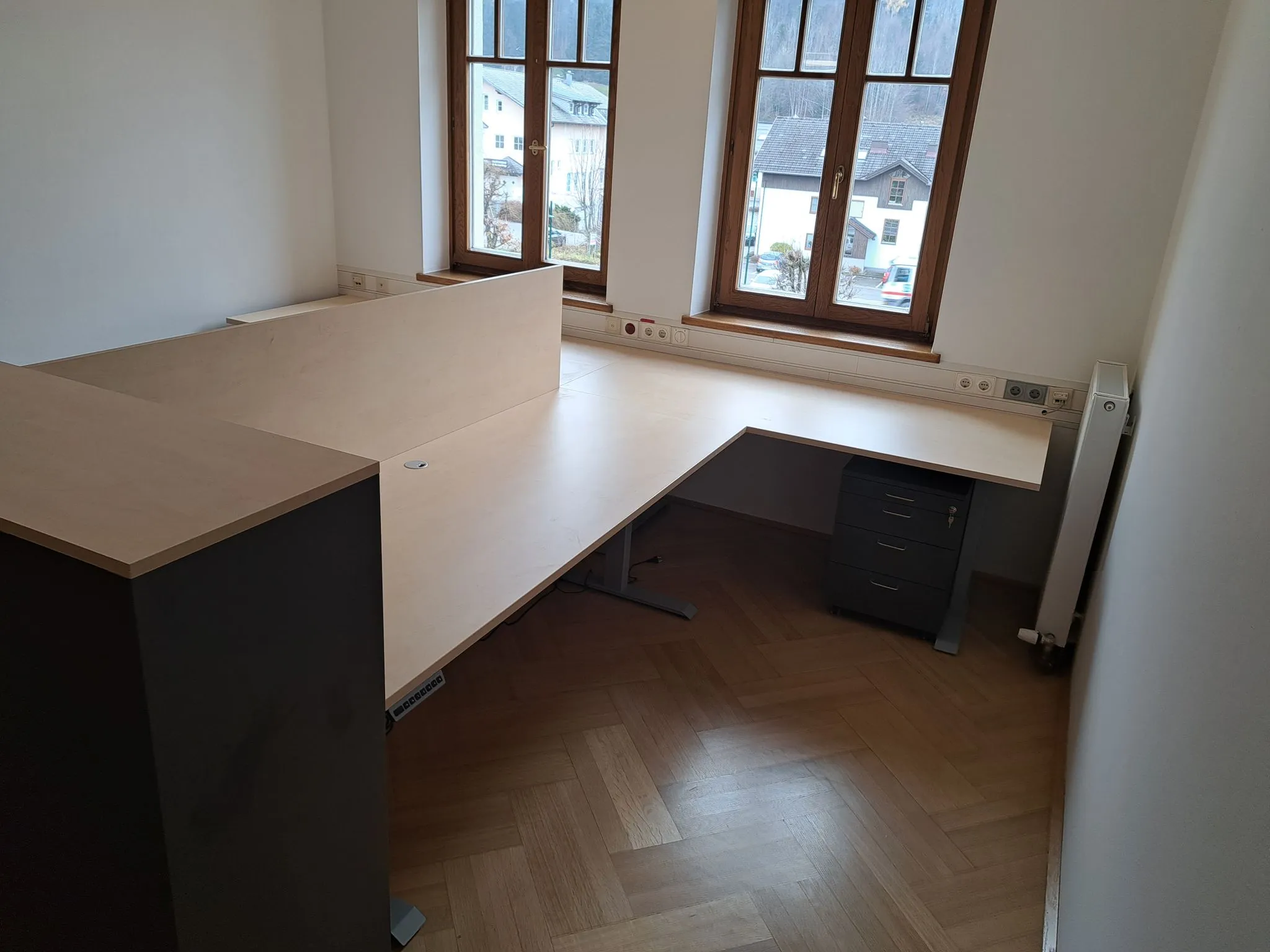 Leerer moderner Eckschreibtisch aus hellem Holz vor zwei Fenstern mit Blick auf Wohnhäuser.