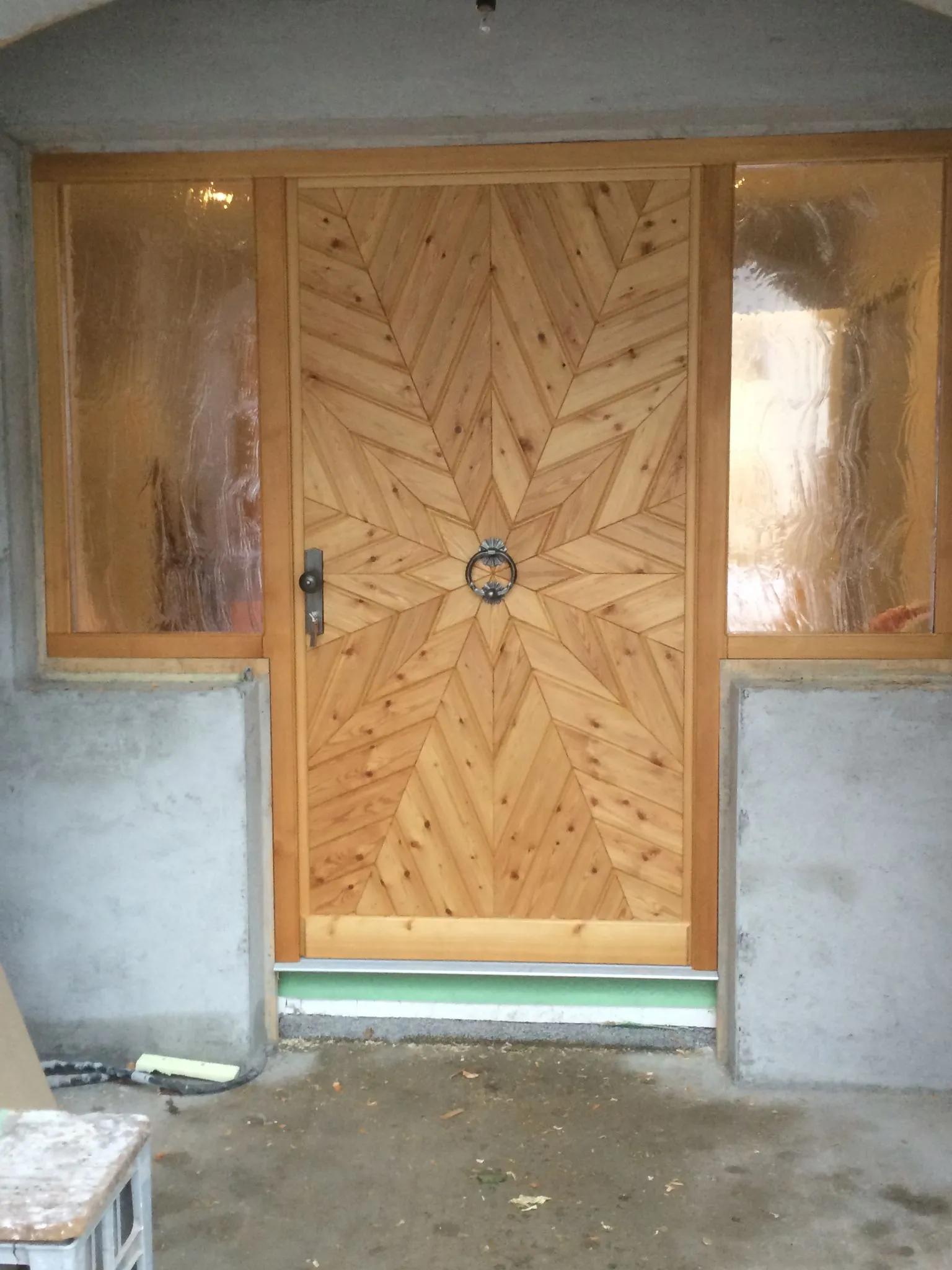 Holztür mit einem sternförmigen Muster aus Holzpaneelen und einem schwarzen Türklopfer, flankiert von zwei undurchsichtigen Fenstern.