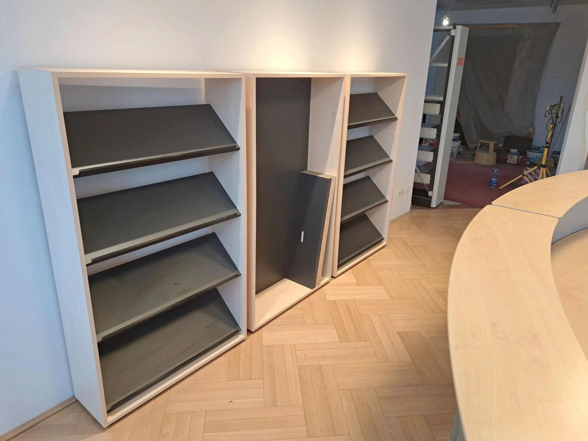 Drei moderne Regale mit schwarzen schrägen Ablagen in einem Raum mit Parkettboden und einem gebogenen Holztisch.