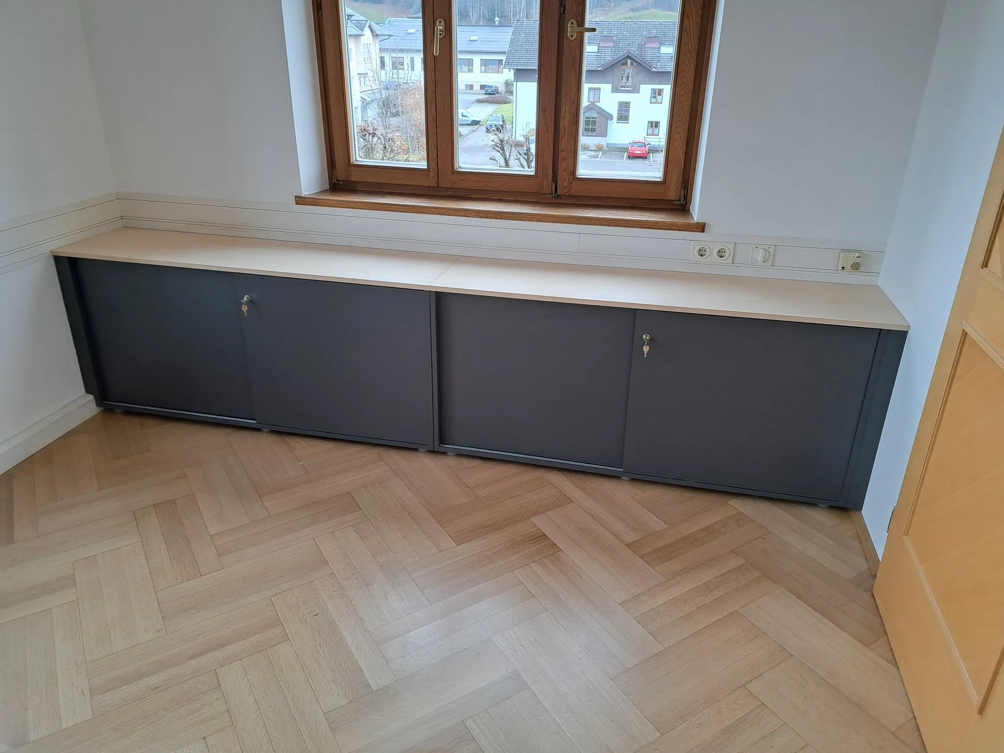 Langes niedriges Sideboard mit schwarzen Türen und heller Holzplatte vor einem Fenster in einem Raum mit Parkettboden.