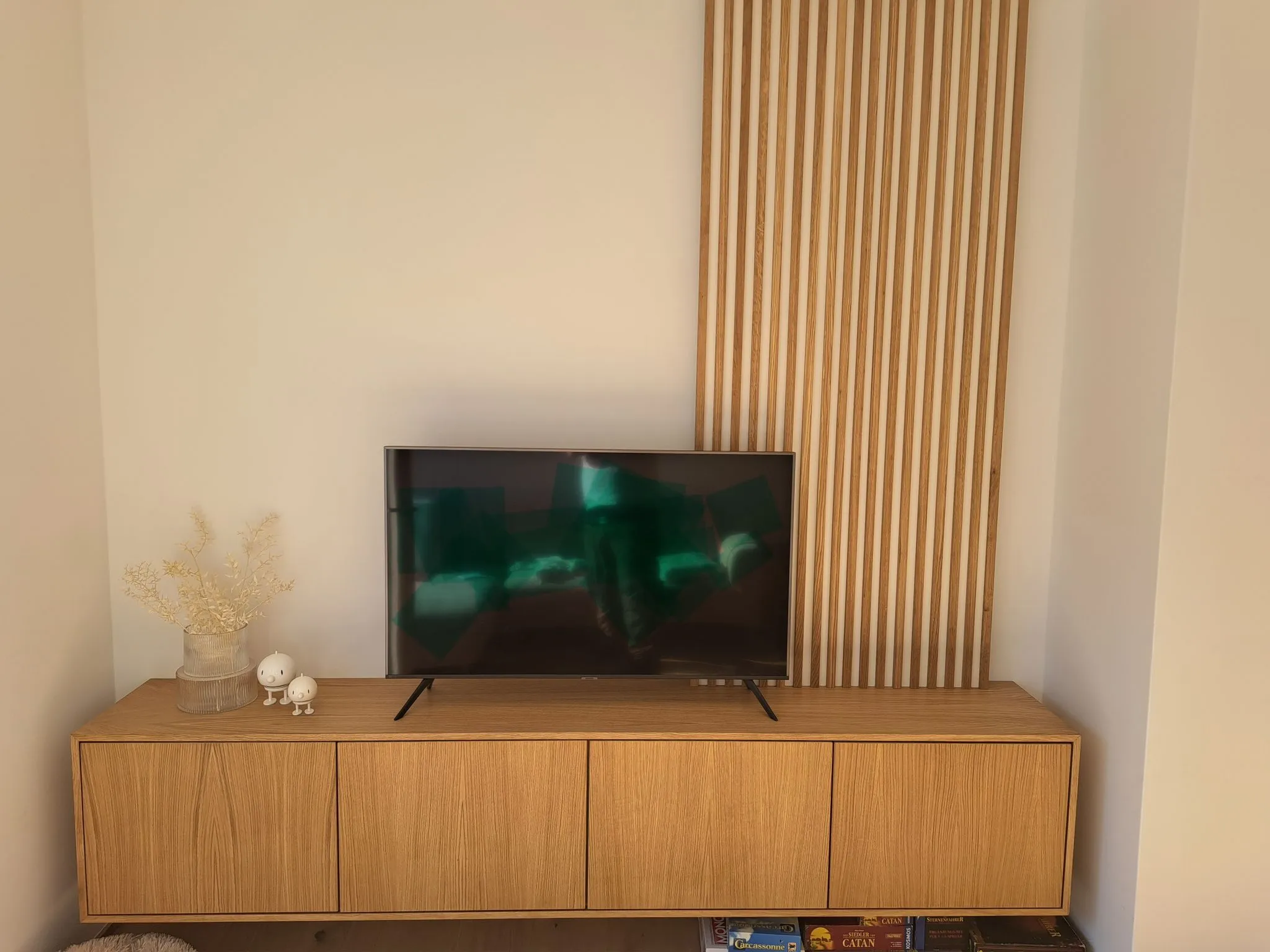 Moderner Wohnzimmerschrank aus Holz mit schwarzem Flachbildfernseher, zwei kleinen weißen Figuren und getrockneten Pflanzen in einem Glasvase.