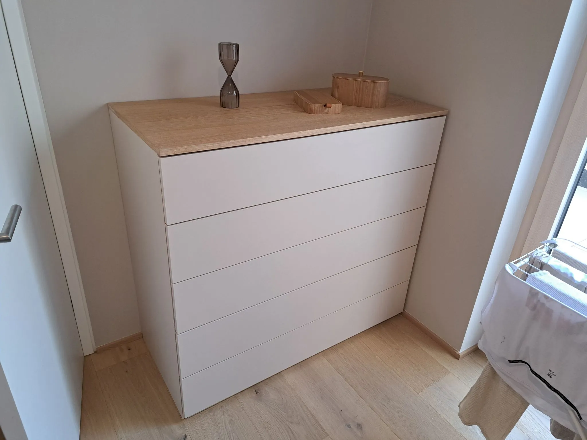 Weißer Kommodenschrank mit fünf Schubladen und einer Holzplatte, darauf eine Sanduhr und zwei kleine Holzbehälter.