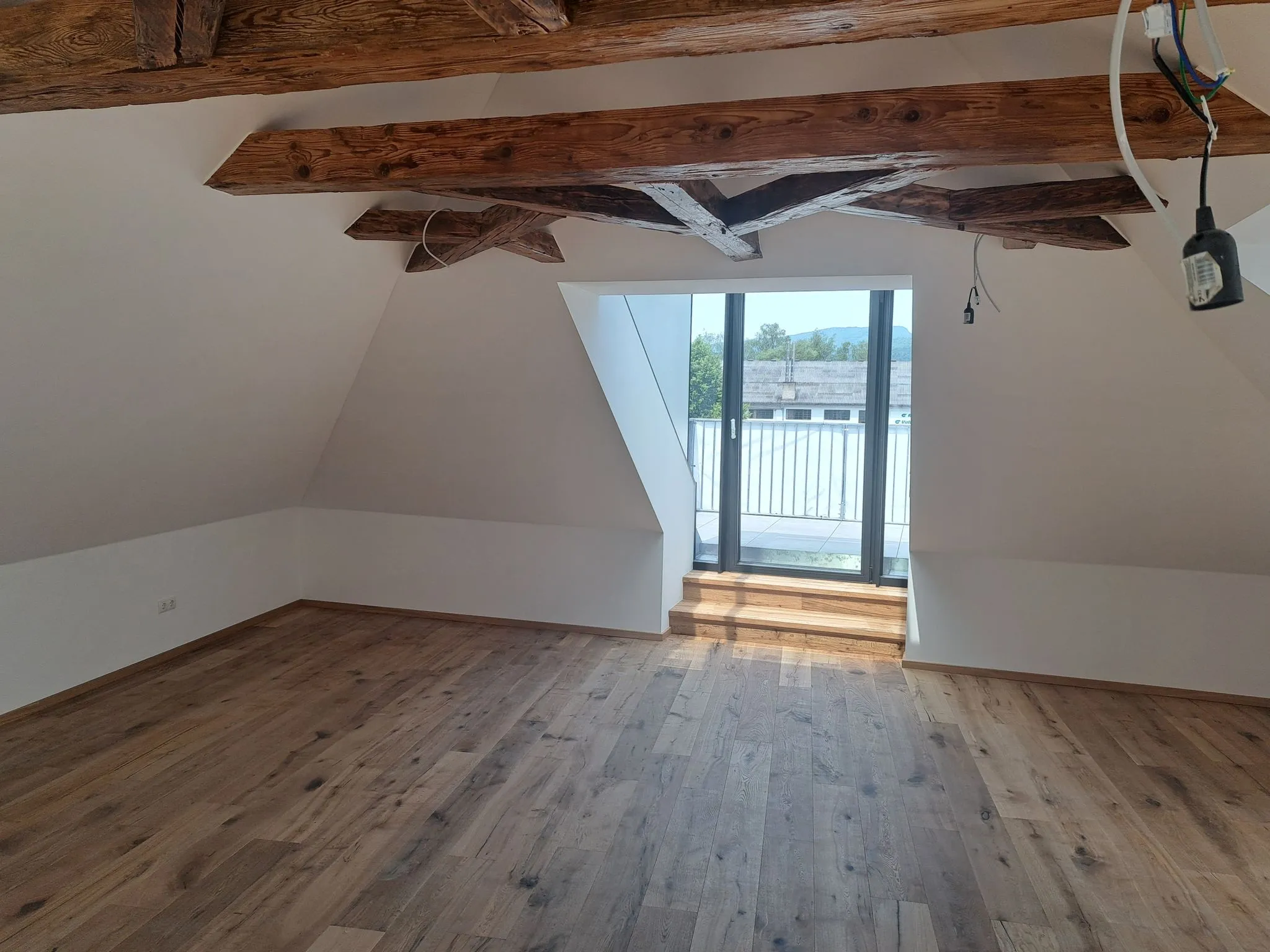 Leerer Dachbodenraum mit hellbraunem Holzboden, weißen Wänden, sichtbaren Holzbalken an der Decke und einer Glastür, die zu einem Balkon führt.