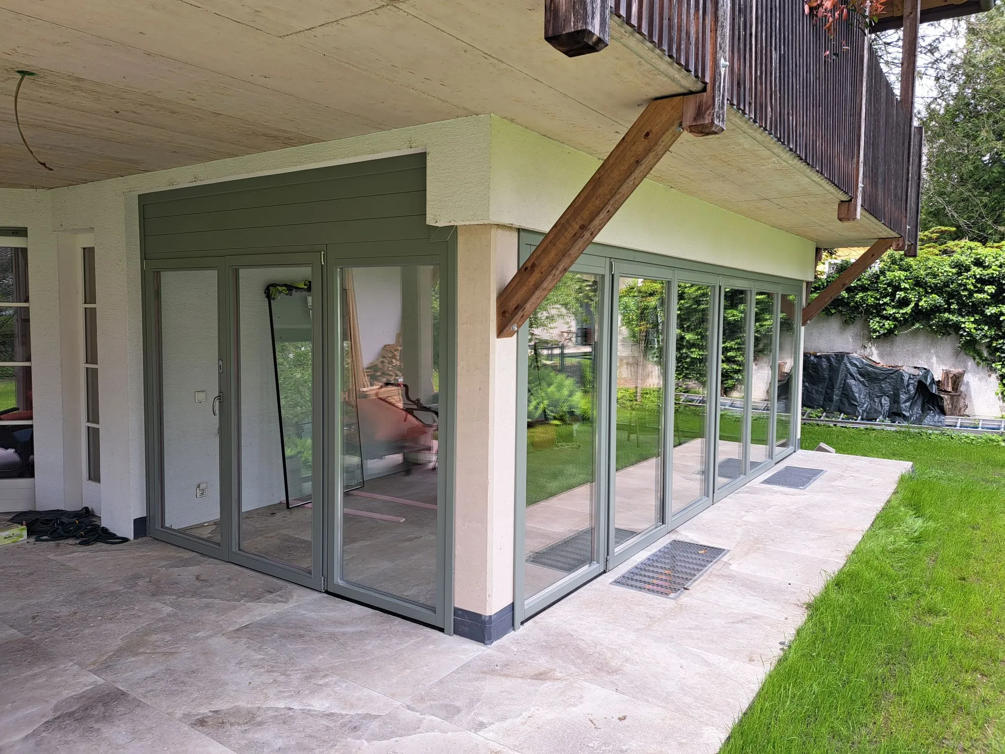 Eckiger verglaster Wintergarten mit grauen Rahmen und großer Terrassenfläche aus beige-farbenen Steinplatten.