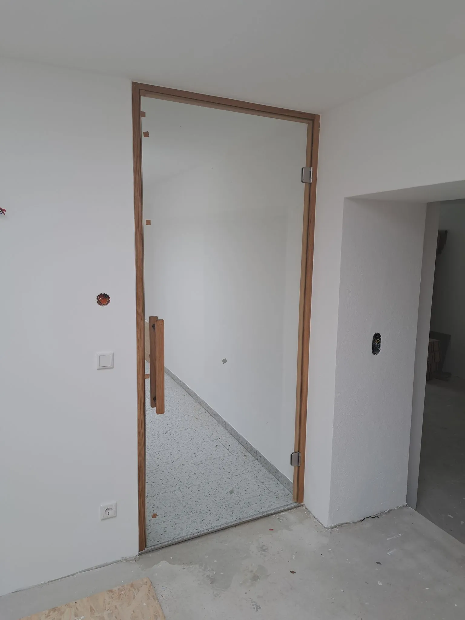 Glastür mit Holzrahmen und Holzgriff in einem weißen, unfertigen Raum mit Fliesenboden im dahinterliegenden Raum.