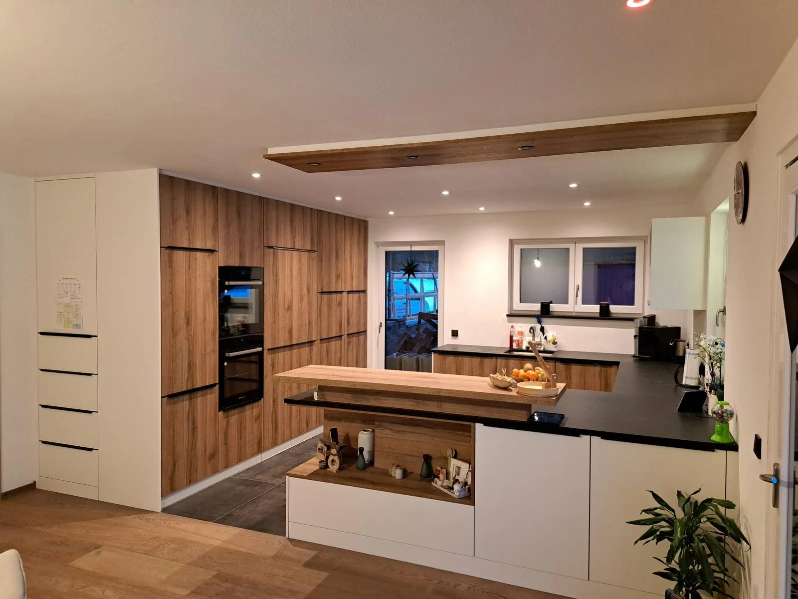 Moderne Wohnküche mit Holzfronten, schwarzen Arbeitsplatten, integrierten Backöfen, einer Kücheninsel mit Holztheke und einer Pflanze in der Ecke.