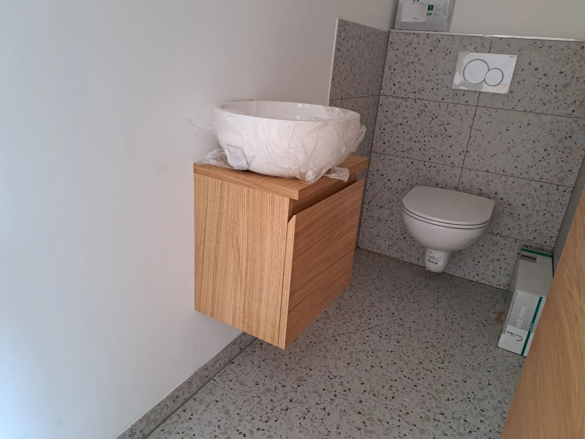 Modernes Badezimmer mit wandhängendem Holzwaschbeckenunterschrank, weißem rundem Waschbecken in Folie und wandhängendem WC vor grauen Fliesen.