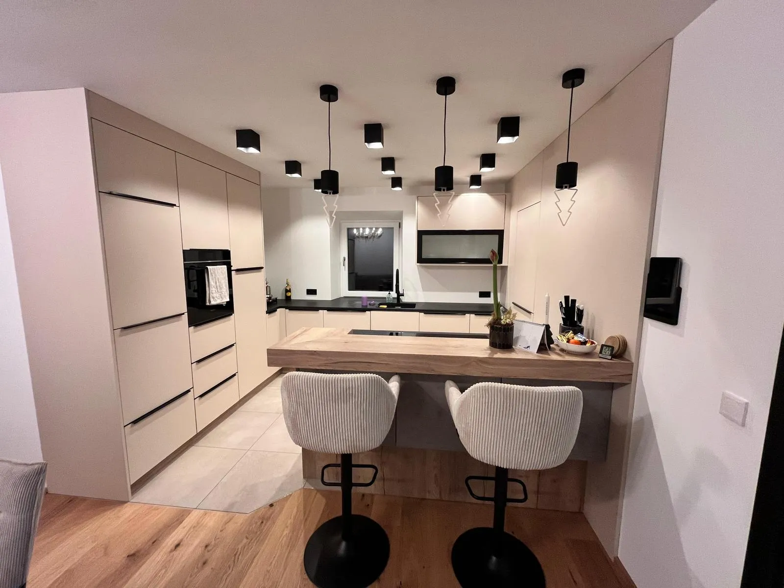 Moderne Küchenzeile mit beigen Schränken, schwarzer Arbeitsplatte, zwei hellen Barhockern und mehreren schwarzen Deckenleuchten.