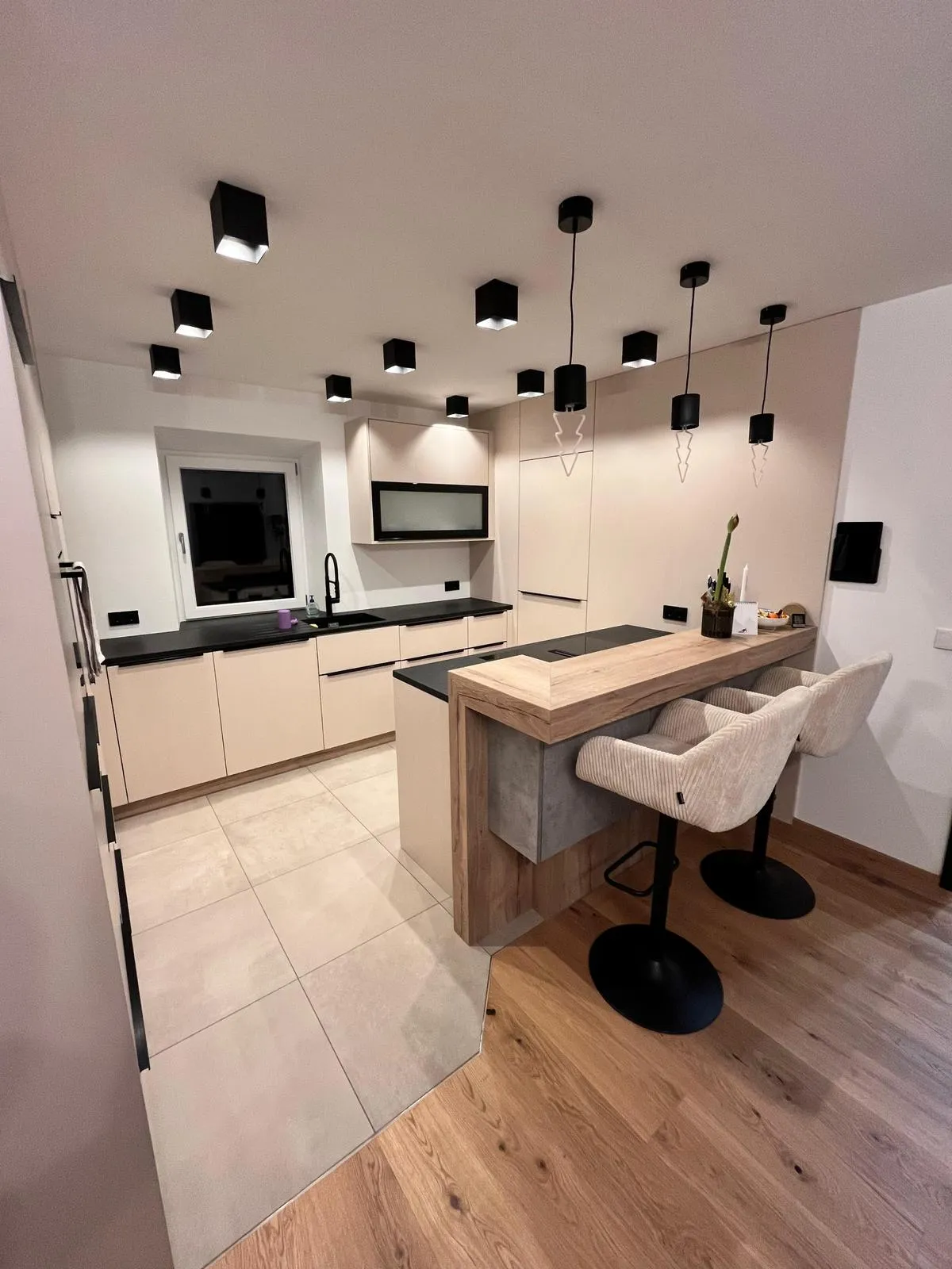 Moderne Küche mit hellen Schränken, schwarzen Arbeitsflächen, zwei gepolsterten Barhockern an einer Holztheke und mehreren schwarzen Deckenleuchten.