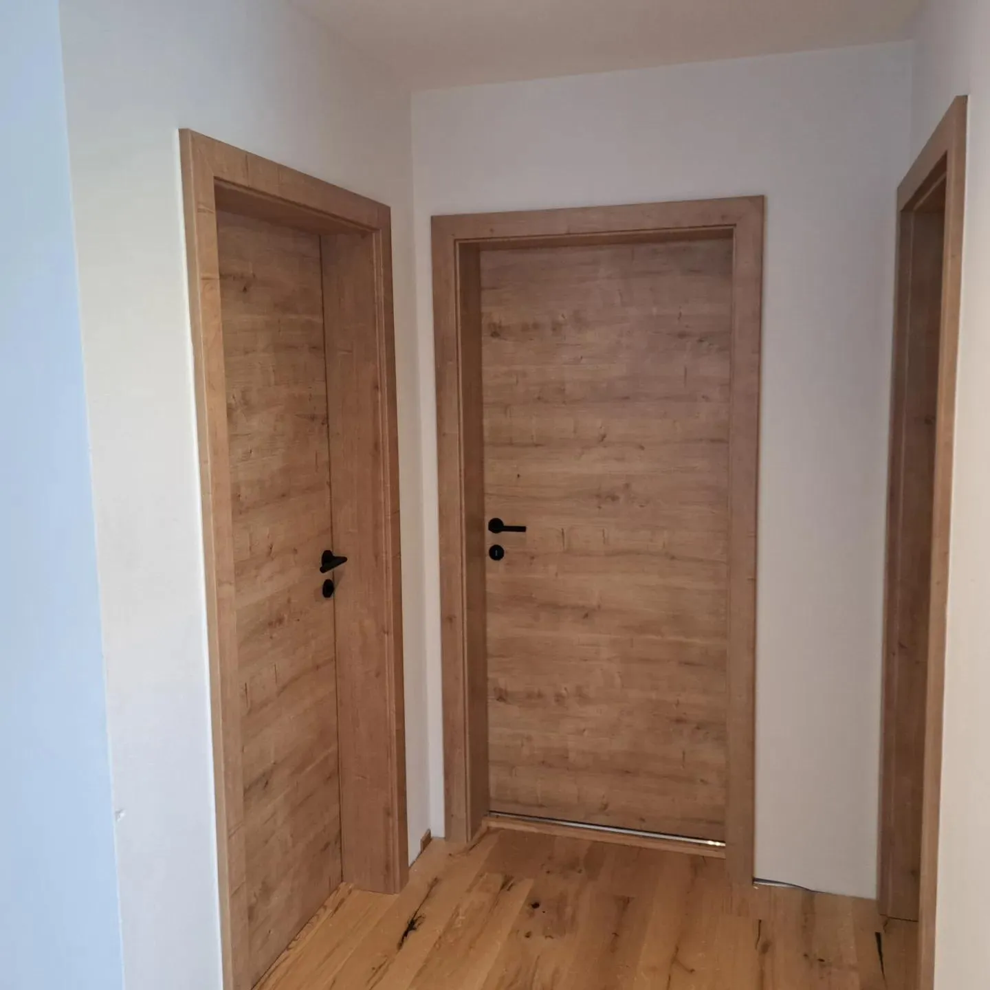 Flur mit drei hellen Holztüren und Holzboden in einem weißen Raum.