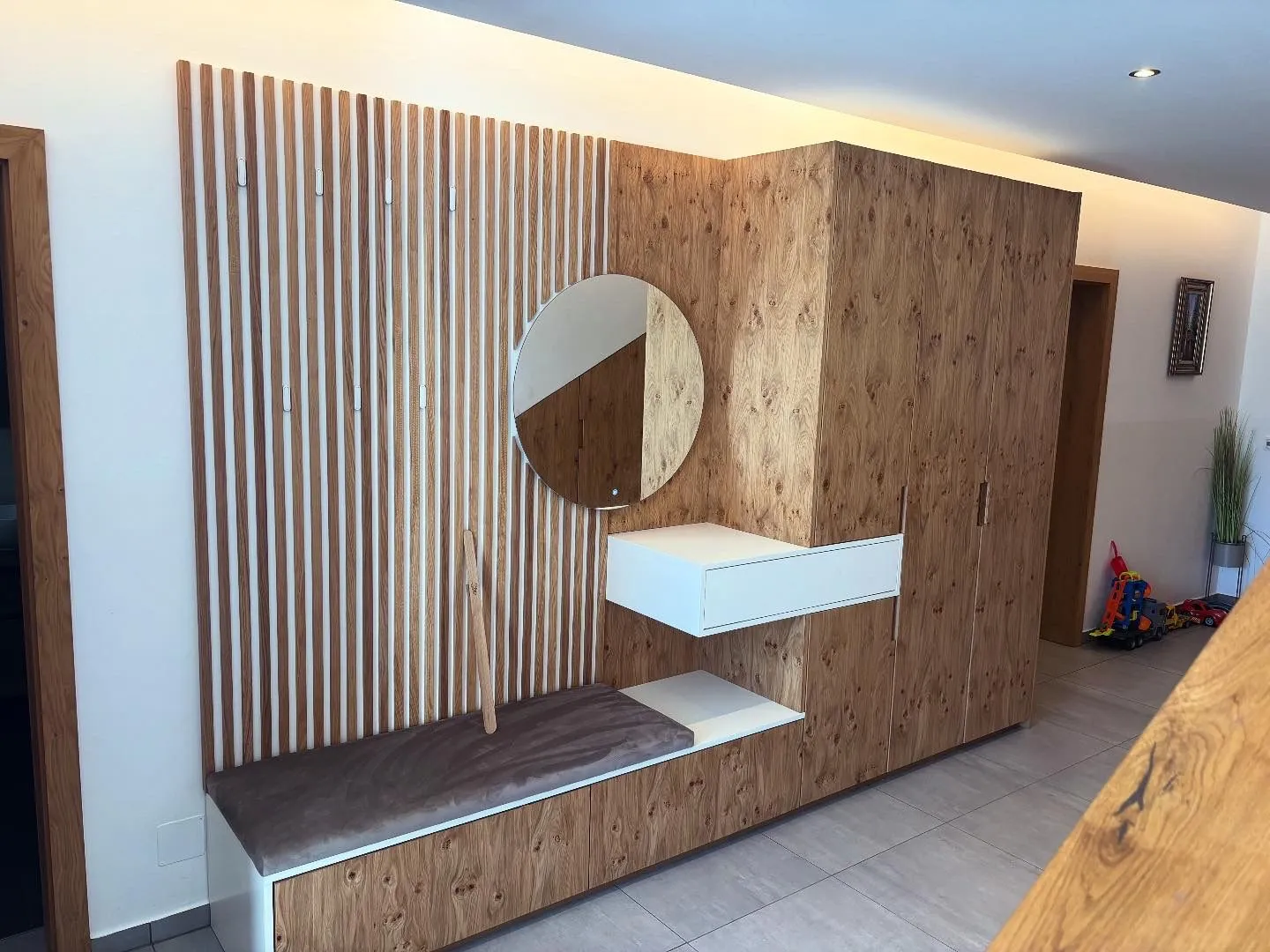 Moderner Holz-Wandbereich mit Sitzbank, Spiegel und Schrank in hellem Flur mit Kinderspielzeug im Hintergrund.